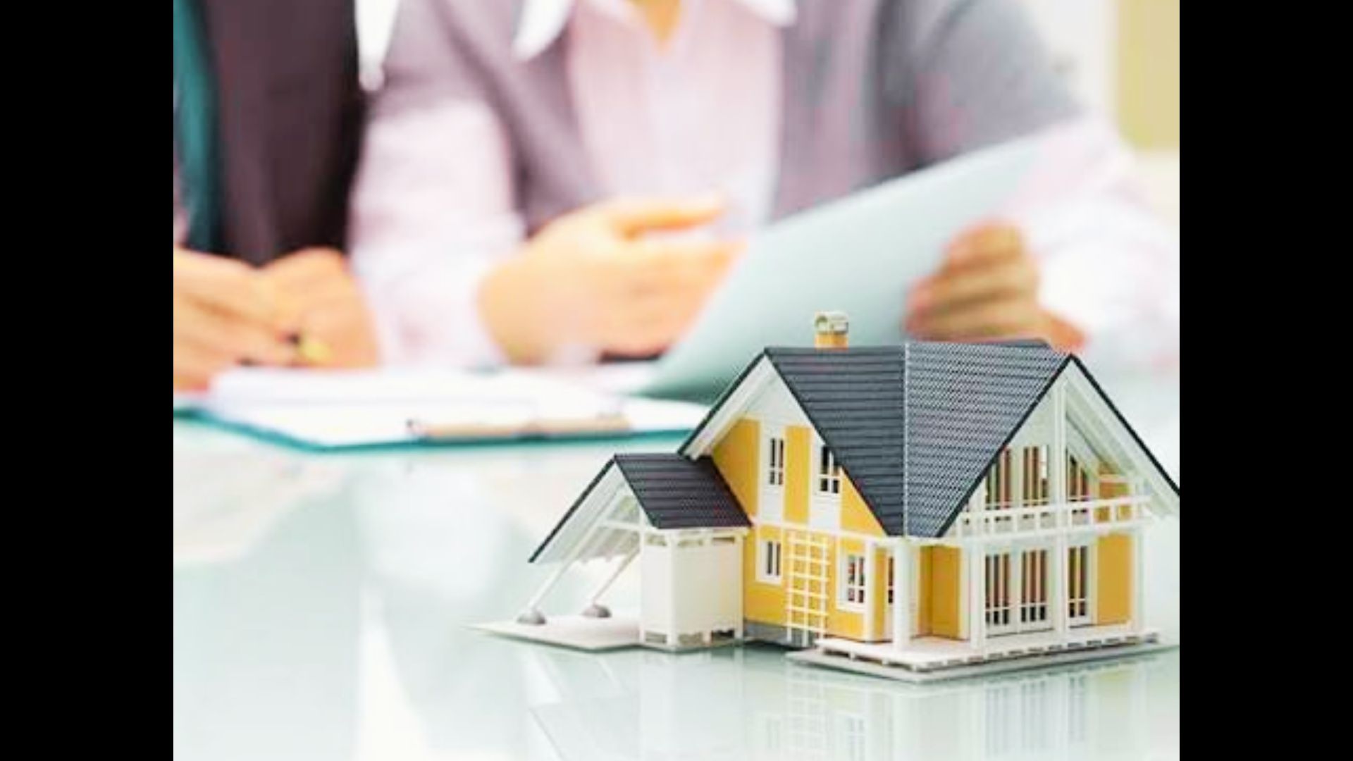 A miniature model house resting on a wooden surface in front of blurred background paperwork.