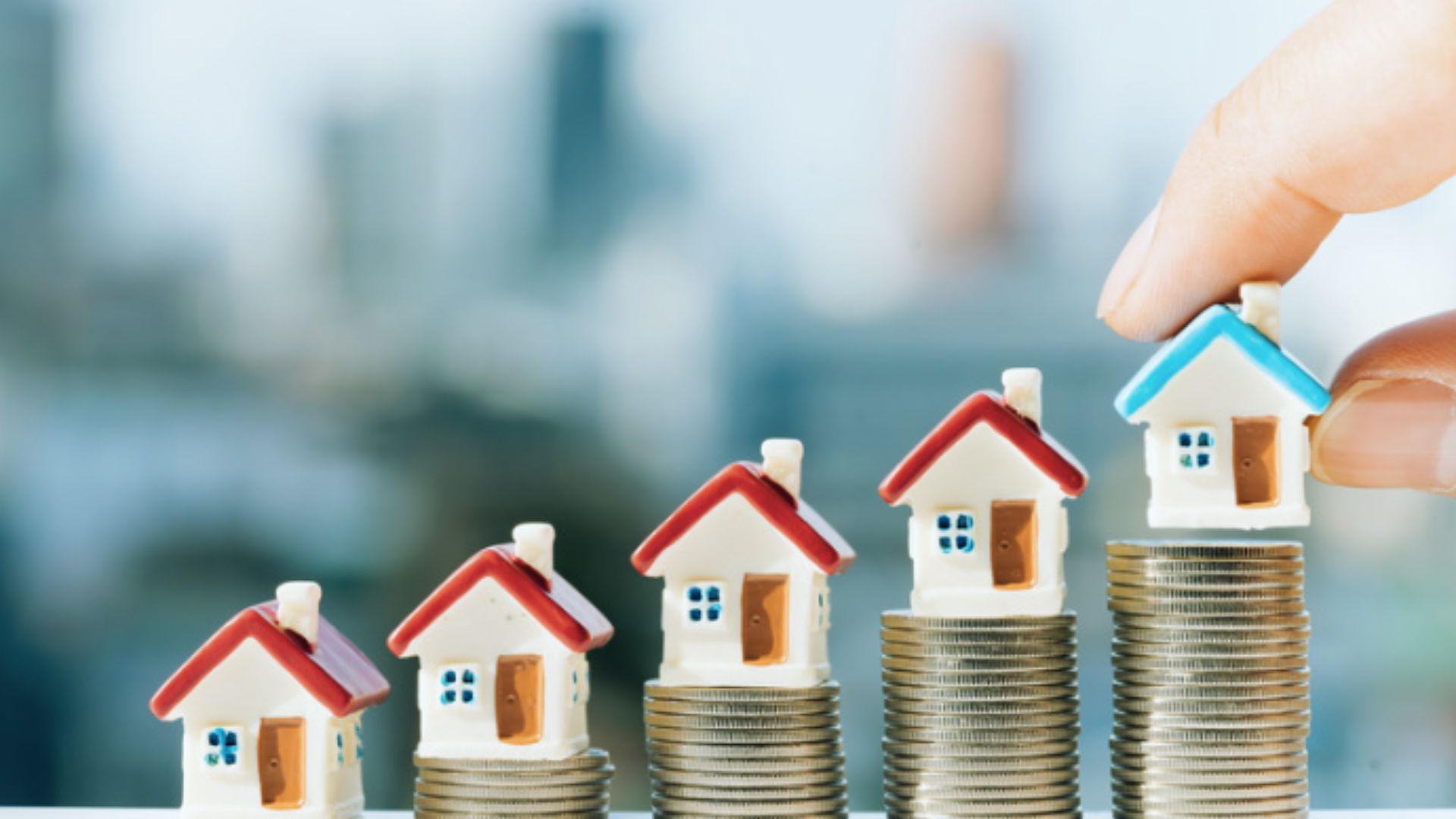 A hand points at a stack of coins topped with miniature houses, symbolizing investment or savings in real estate.