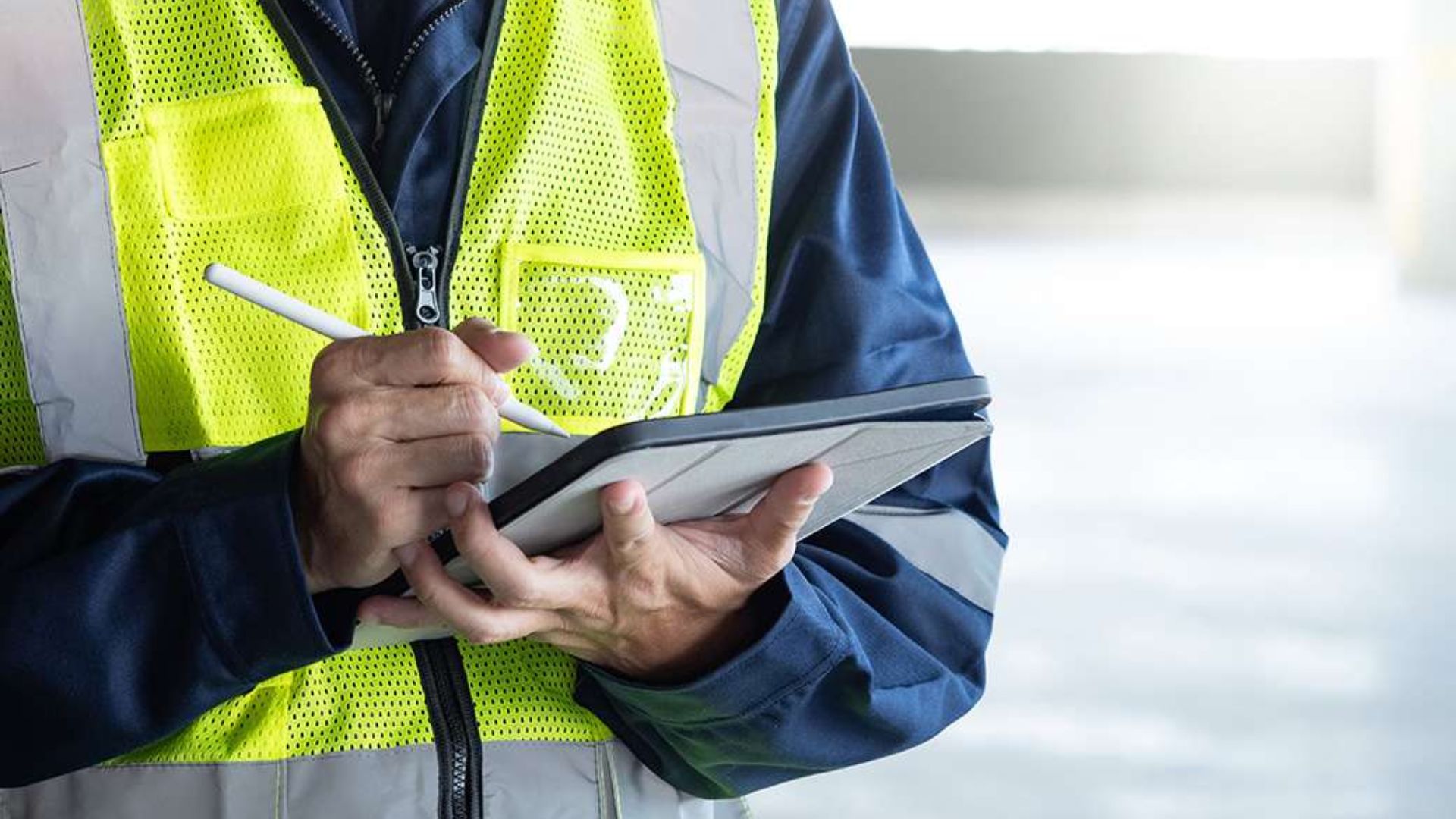 A man in a safety vest writes on a tablet with a pen, focused on his task in a work environment.