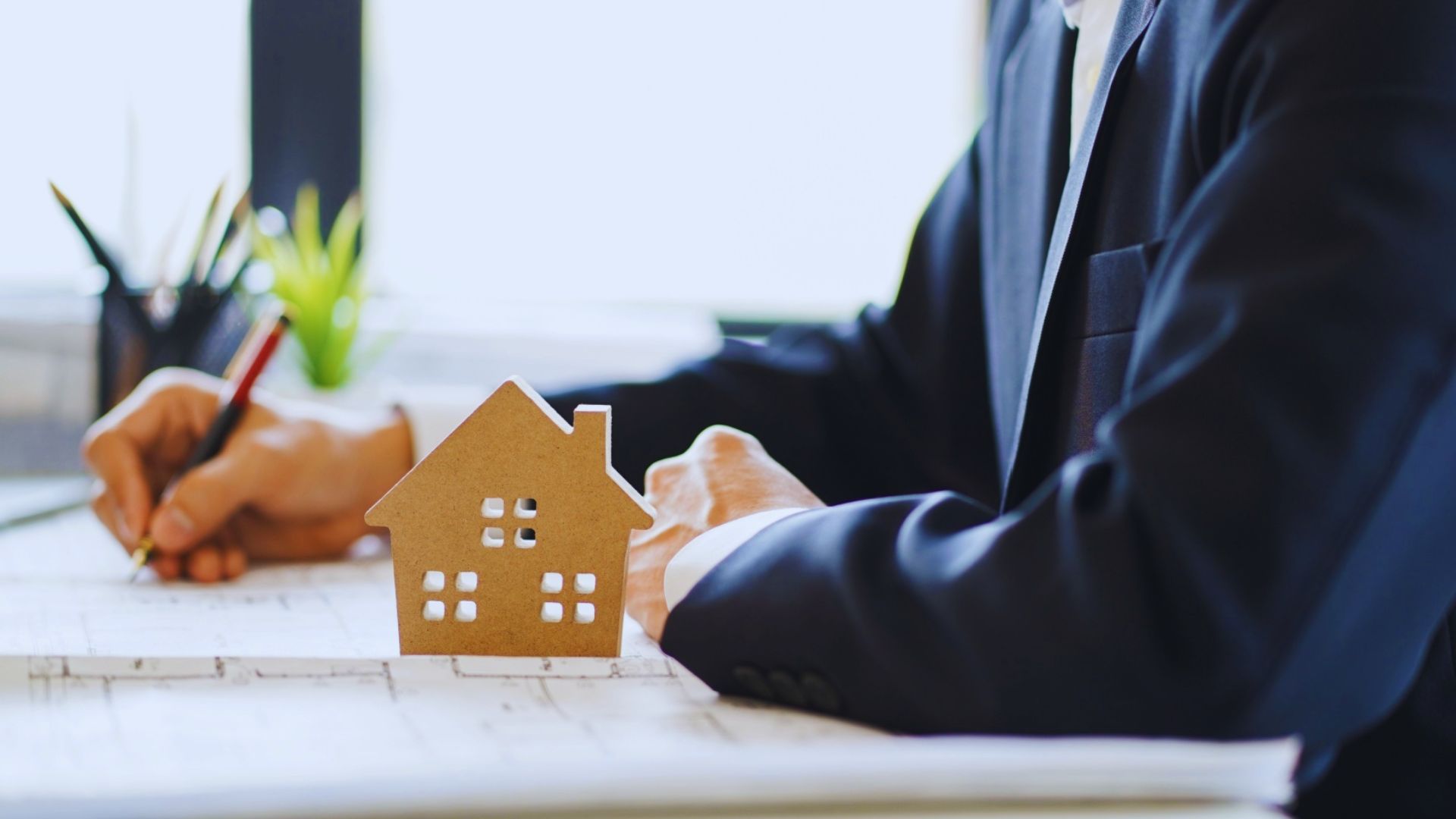 Man in a suit and tie sitting at a desk, examining a small house model with a focused expression.