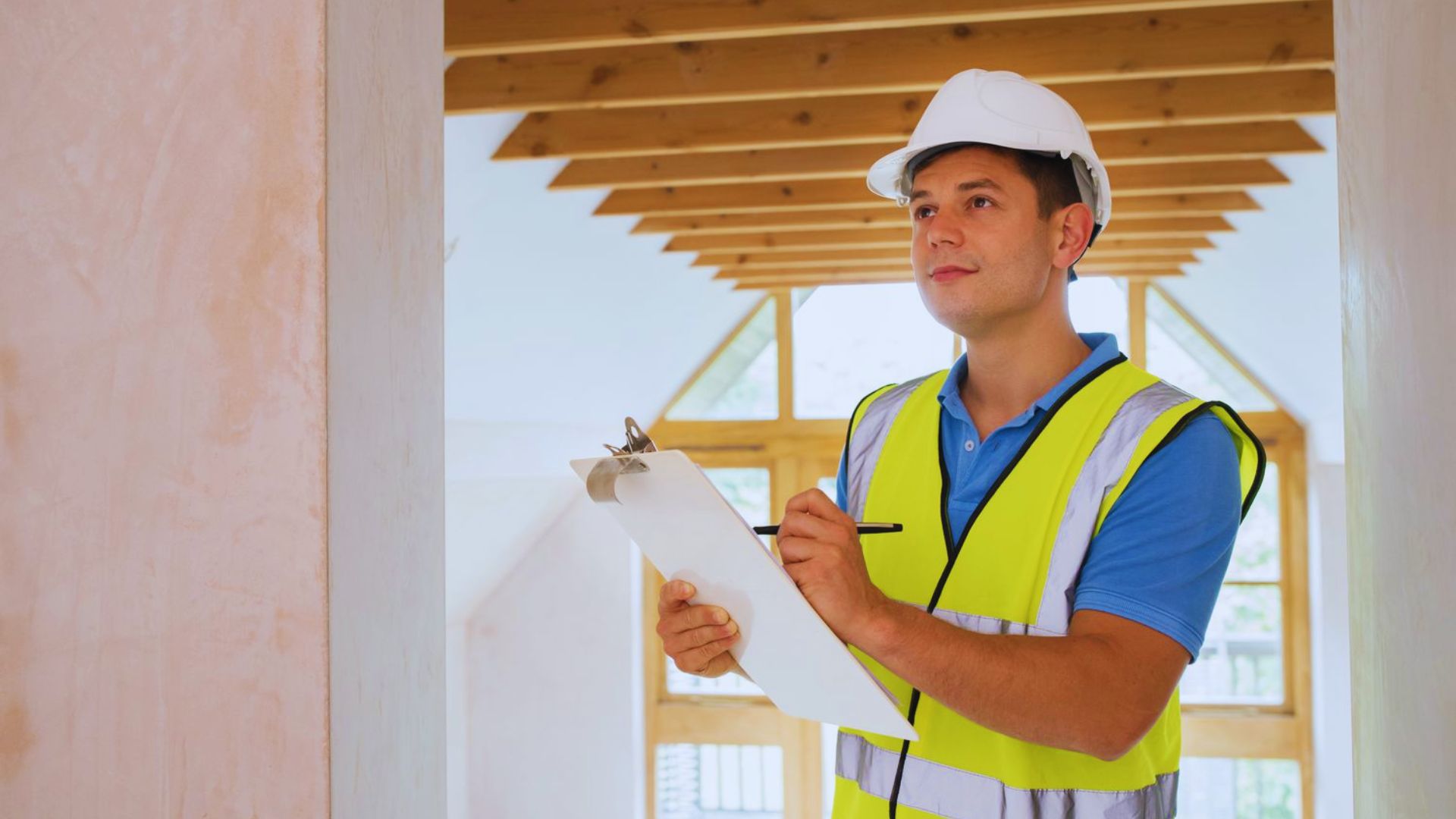 A man wearing a hard hat and safety vest holds a clipboard, standing in a construction environment.