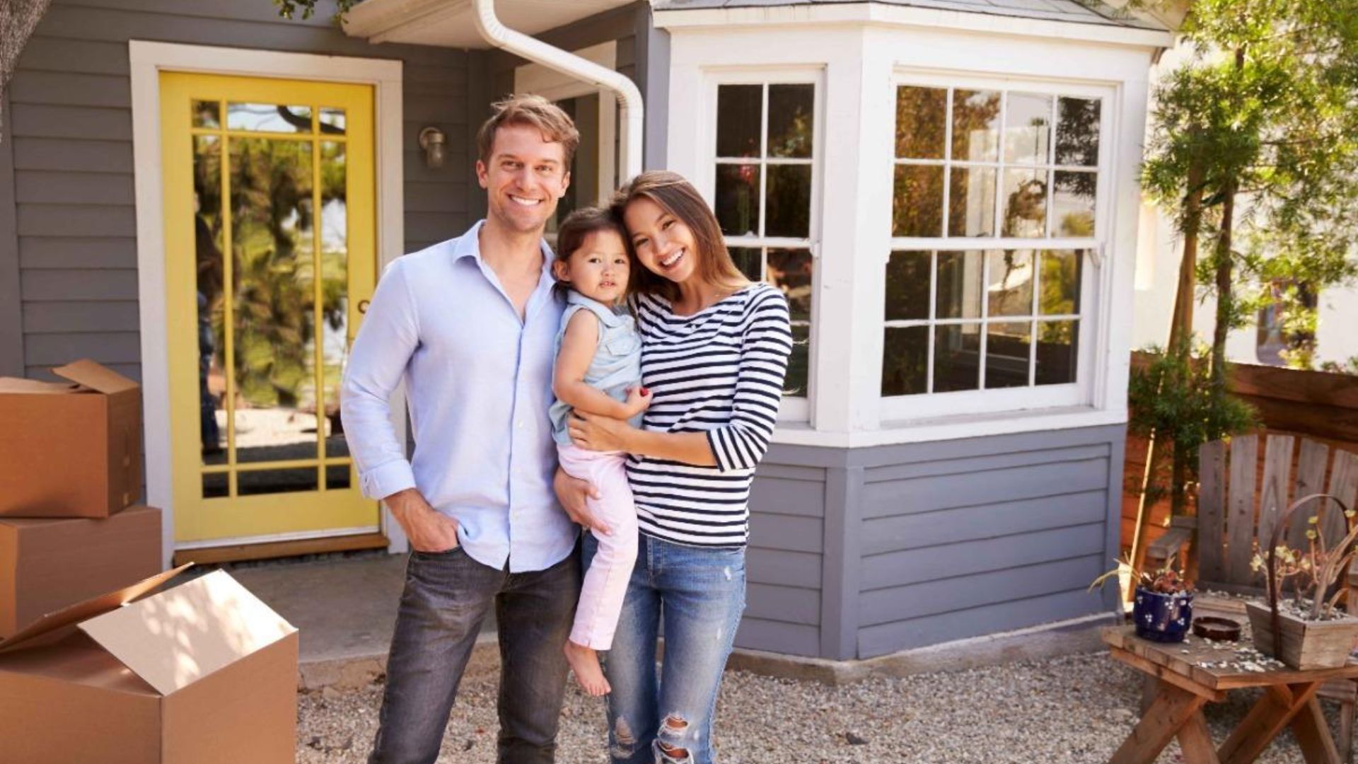 A man and woman stand in front of a house, holding a child together, smiling at the camera.