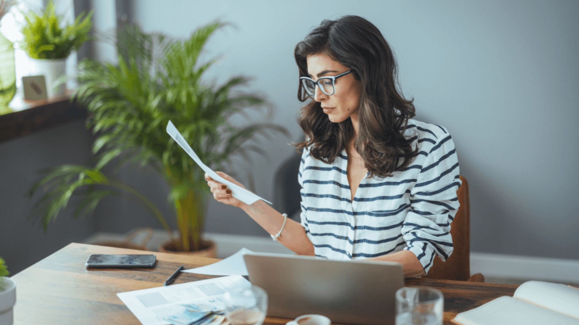 A woman wearing glasses reads a document on her laptop, focused on the screen in a well-lit environment.