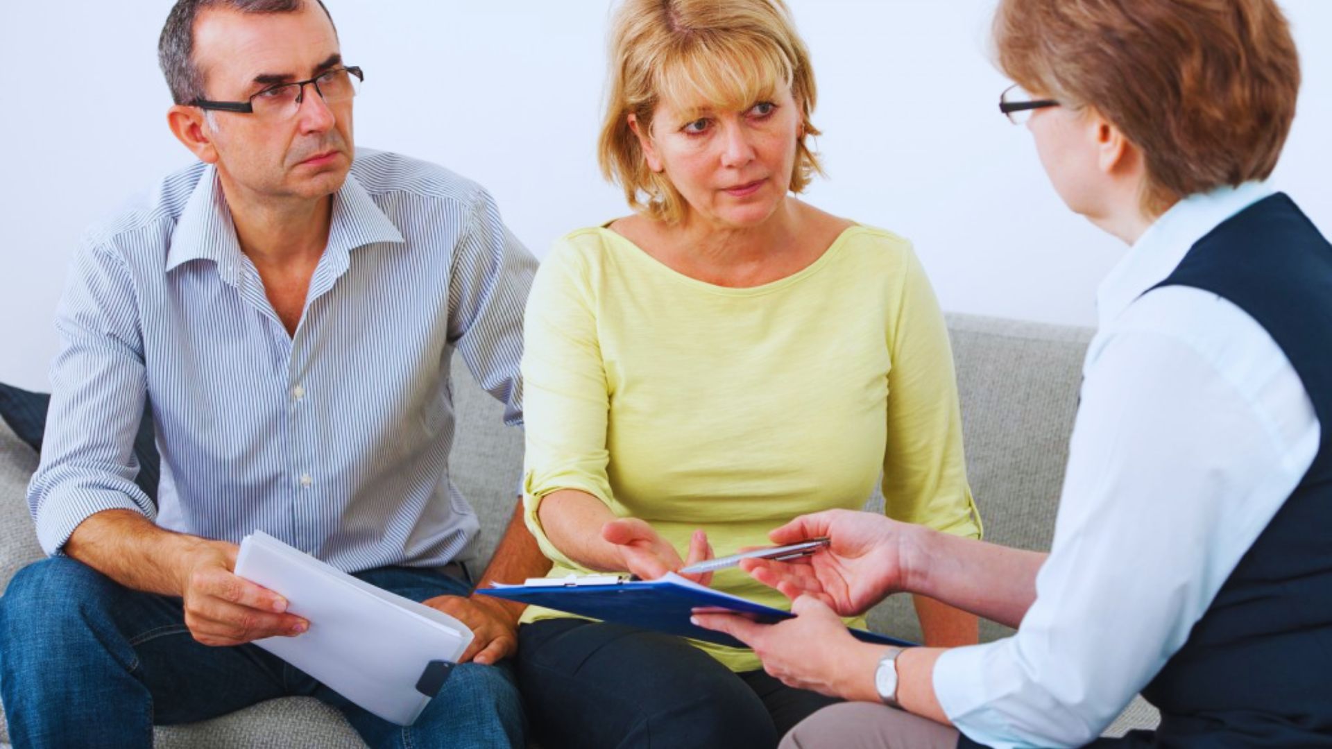 A man and woman sit on a couch; the man holds a clipboard, appearing to discuss something with the woman.