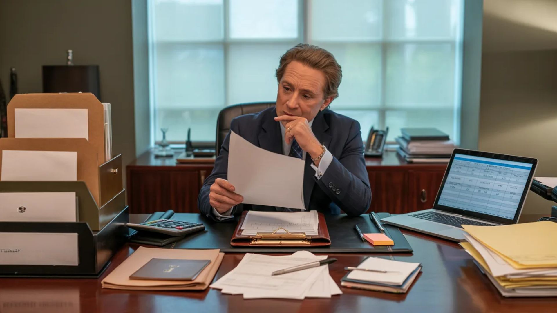 A man in a suit sits at a desk, surrounded by papers and a laptop, focused on his work.