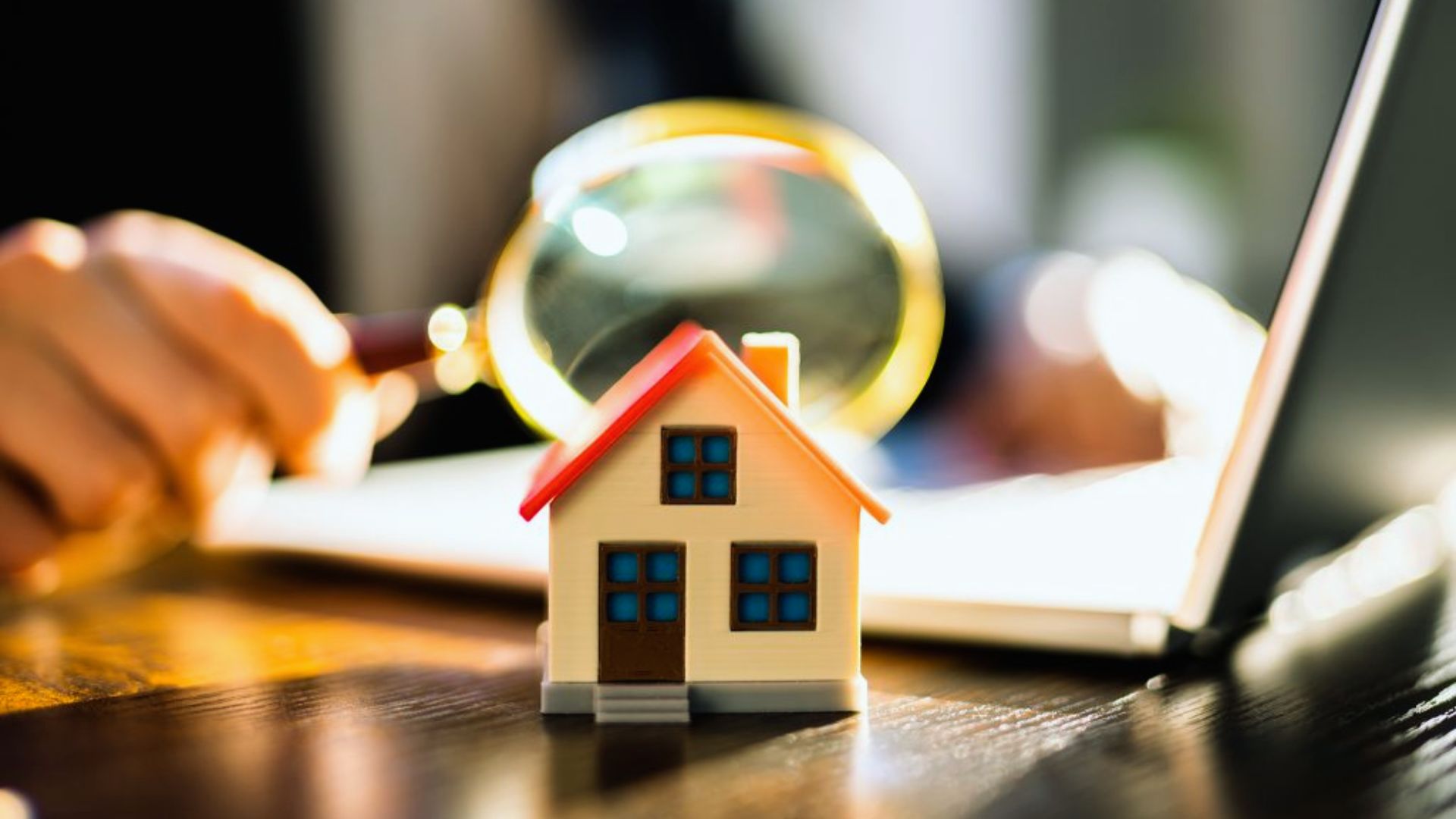 A person's hand holds a house model while a magnifying glass examines it closely.
