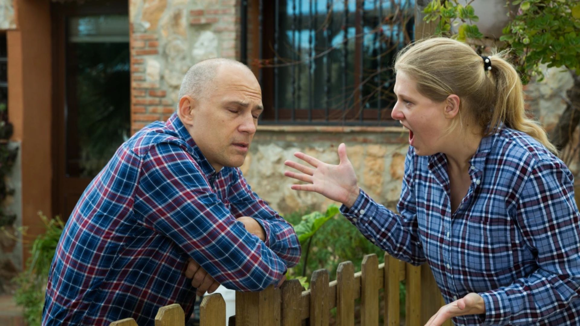 A man and woman are chatting casually while resting on a fence, surrounded by greenery in a bright outdoor environment.