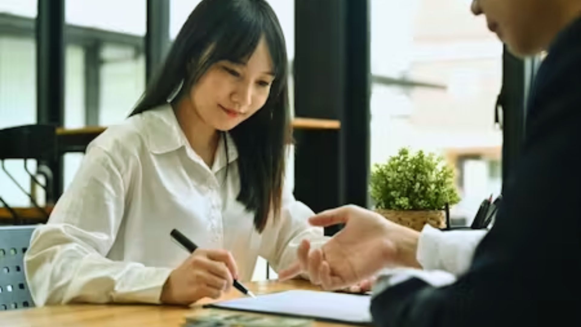Asian woman signing documents with a man in a modern office setting, both focused on the paperwork.
