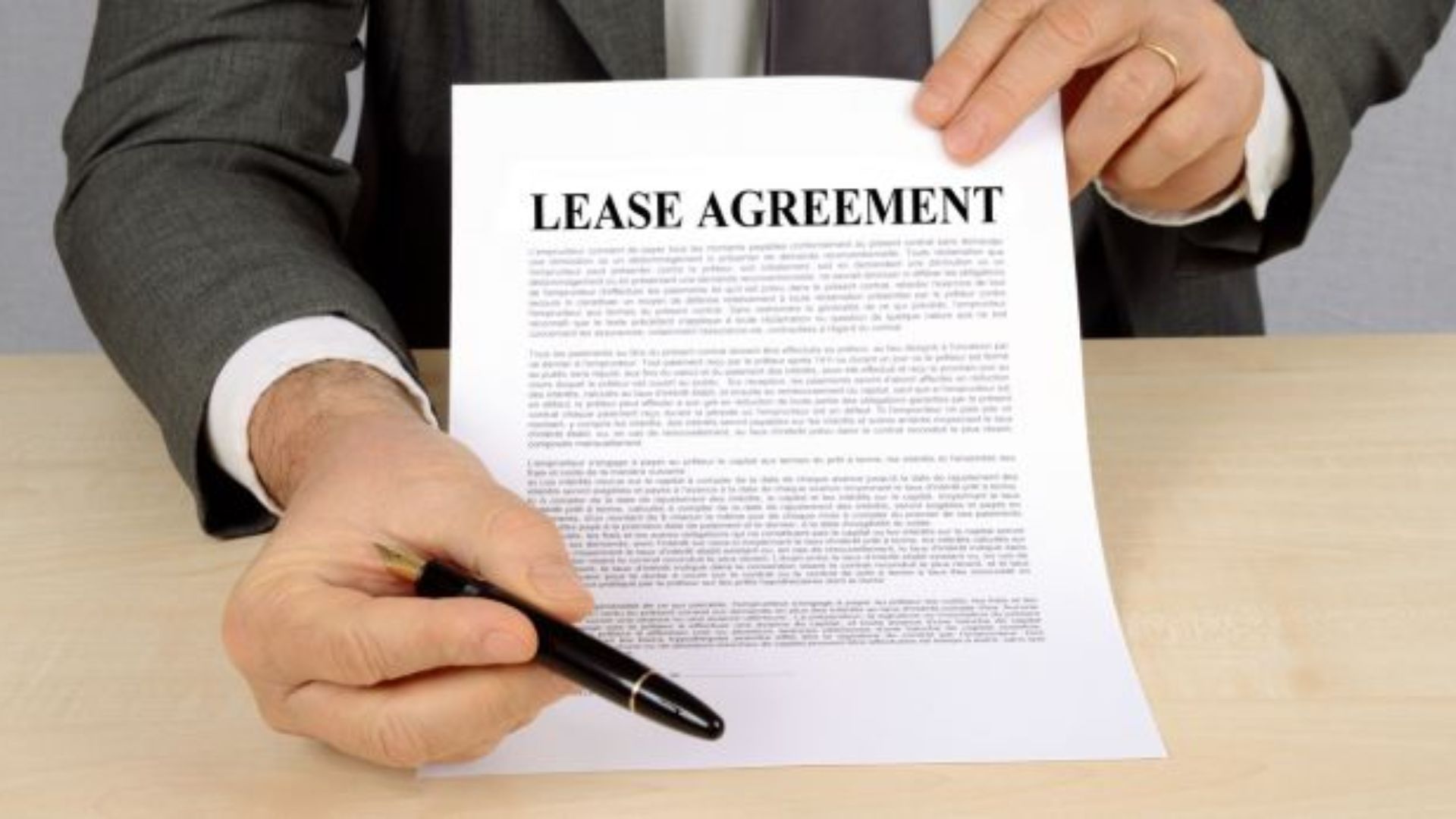A person in a suit holding a lease agreement across a table, offering a pen. The scene conveys a formal and businesslike tone.