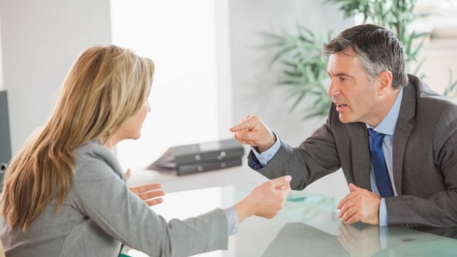A man and woman in business attire are engaged in an intense discussion at a table. Both are leaning forward, gesturing with their hands. The setting is an office.