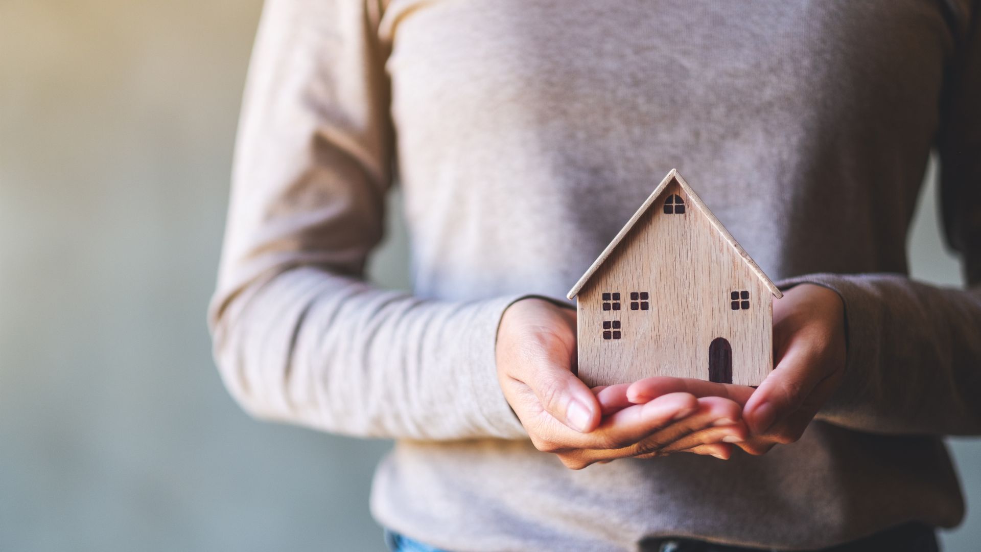 A person in a beige sweater holds a small wooden house model with both hands. The image conveys warmth, care, and the concept of homeownership.