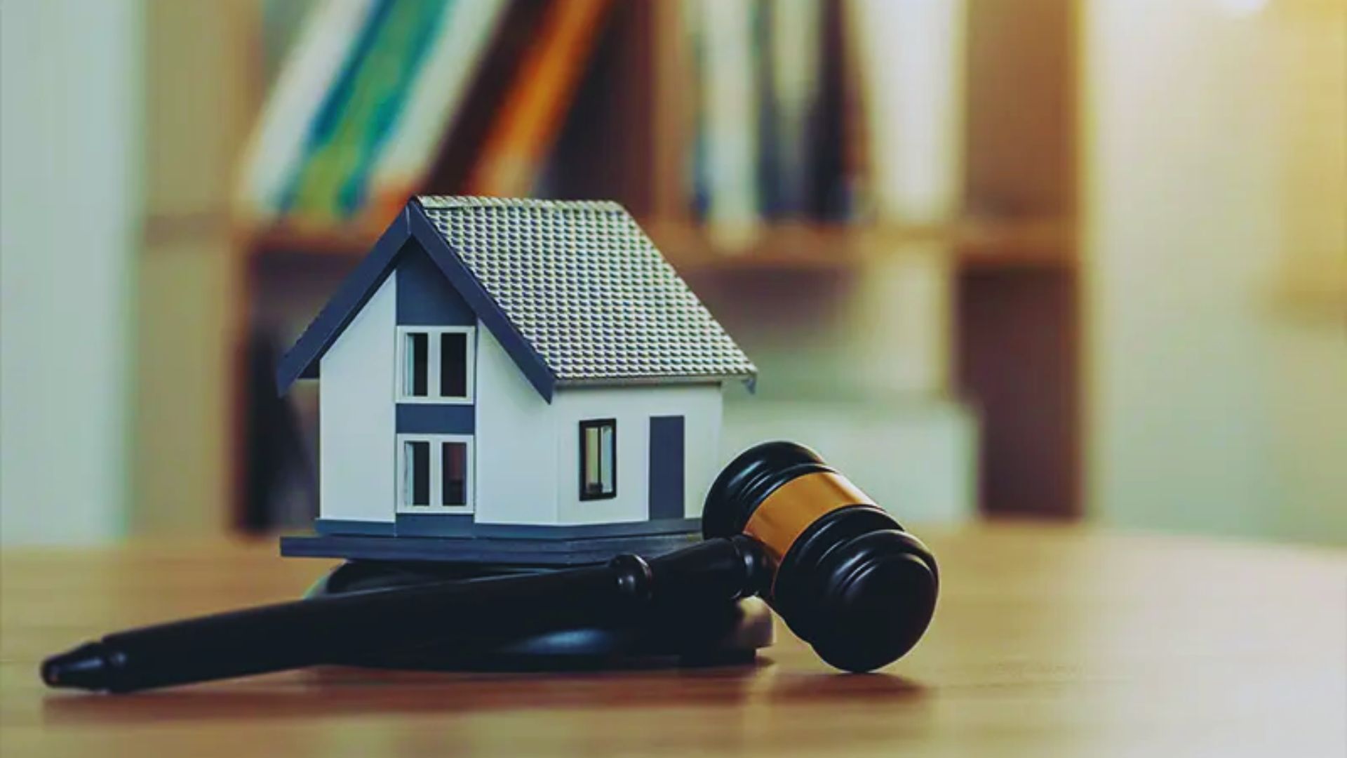 A small model house sits on a wooden table beside a judge's gavel, with blurred bookshelves in the background, suggesting themes of real estate law.