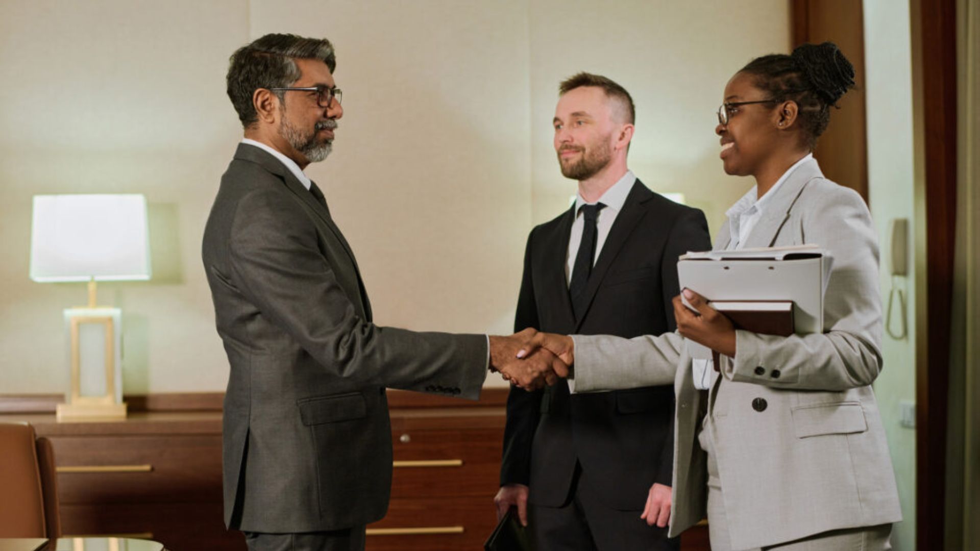 Two business professionals, a man and a woman, shake hands in a formal office setting, while another man stands smiling beside them. All wear suits, conveying professionalism.