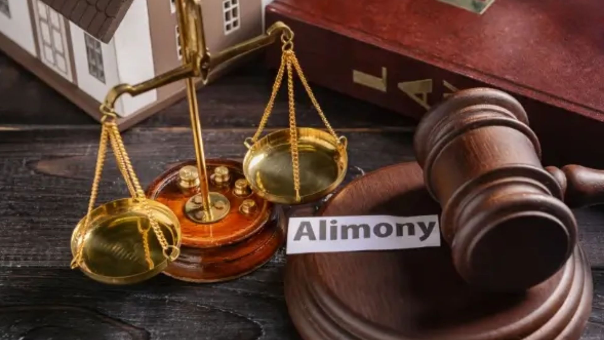 A wooden gavel rests beside golden scales of justice, with a house model and a legal book in the background. A paper labeled "Alimony" is in the scene.