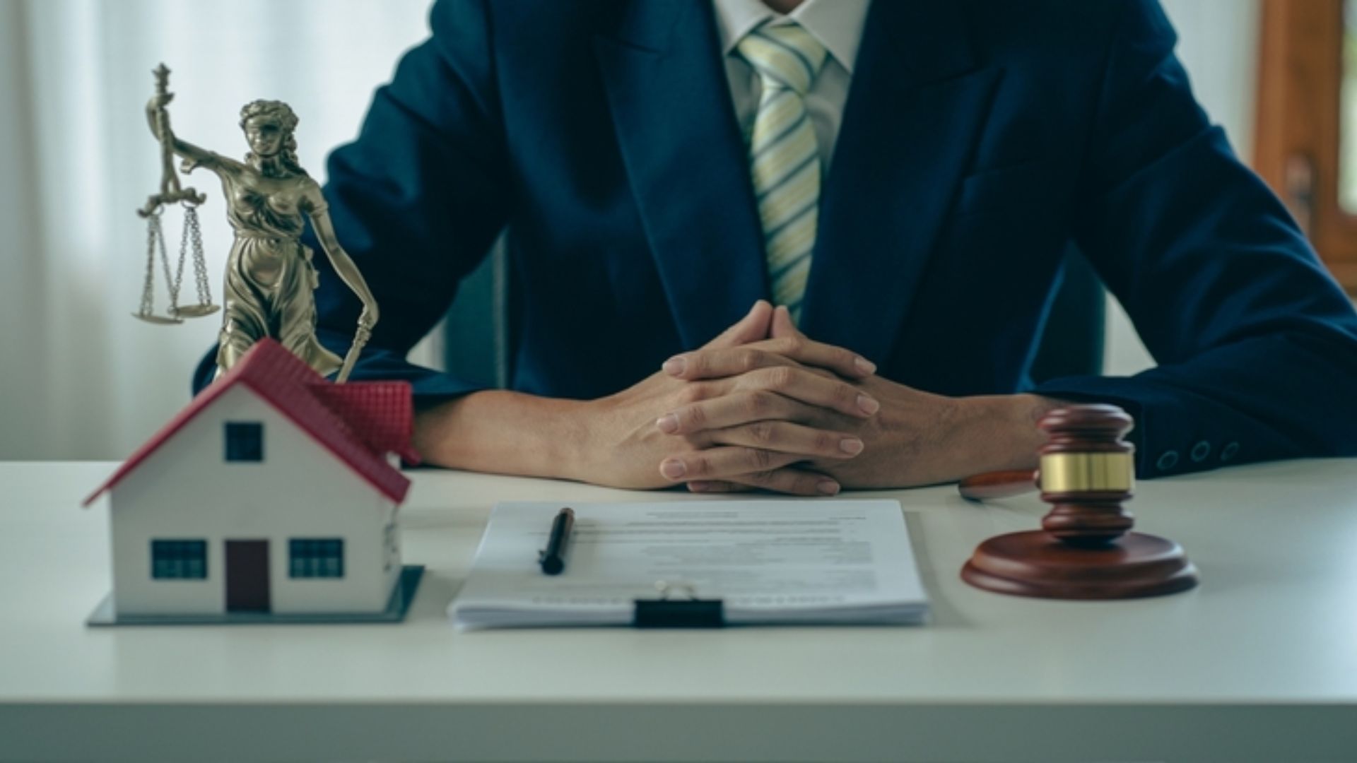 A suited individual sits at a desk with folded hands. In front, there's a small house model, Lady Justice statue, a document, and a gavel, implying legal or real estate matters.