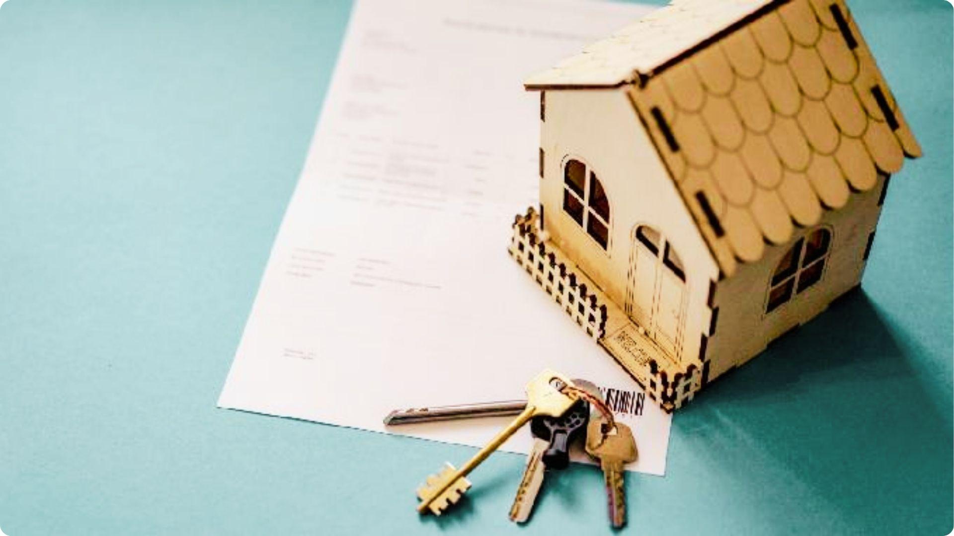 House keys lie on paperwork next to a wooden model home on a teal surface.
