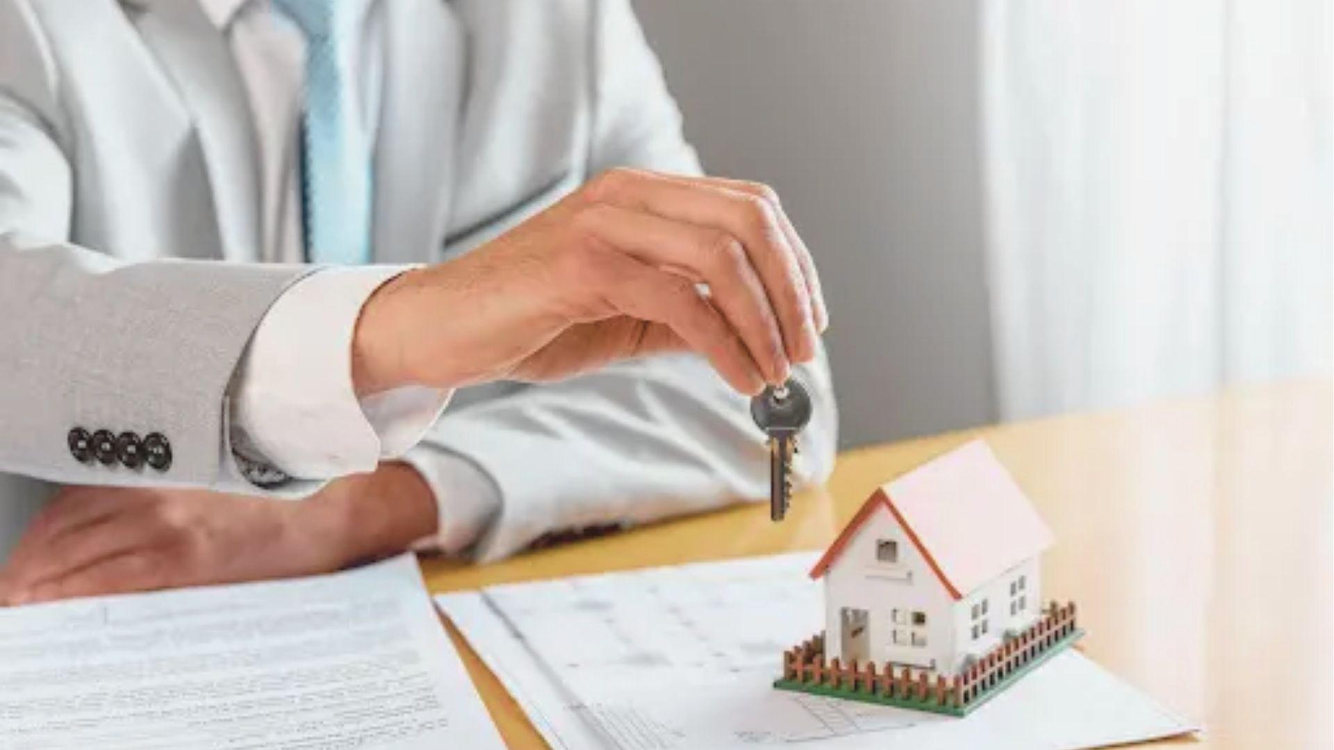 A person in a light gray suit holds house keys over a small model home and legal documents on a desk.