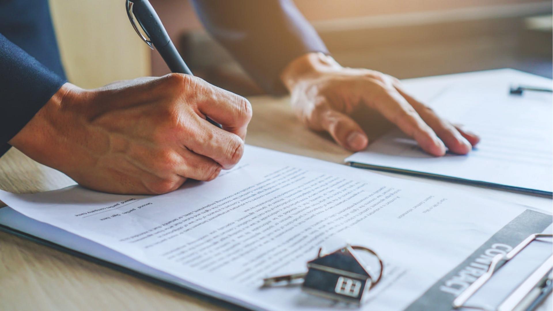 Person signs legal contract on clipboard with house-shaped keychain resting on document, indicating real estate ownership transfer.