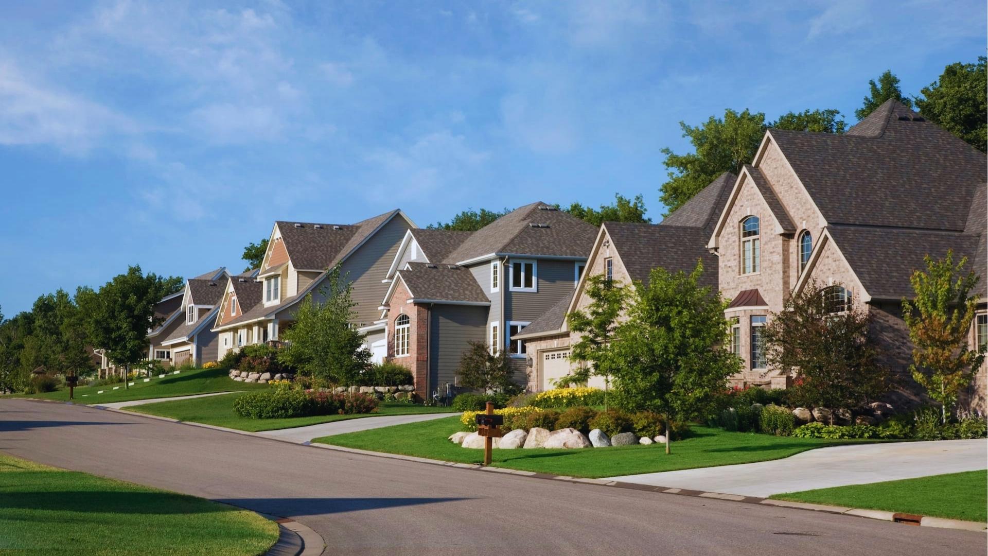 Upscale suburban neighborhood with large two-story brick and siding homes, manicured lawns, mailboxes, and trees under blue sky.