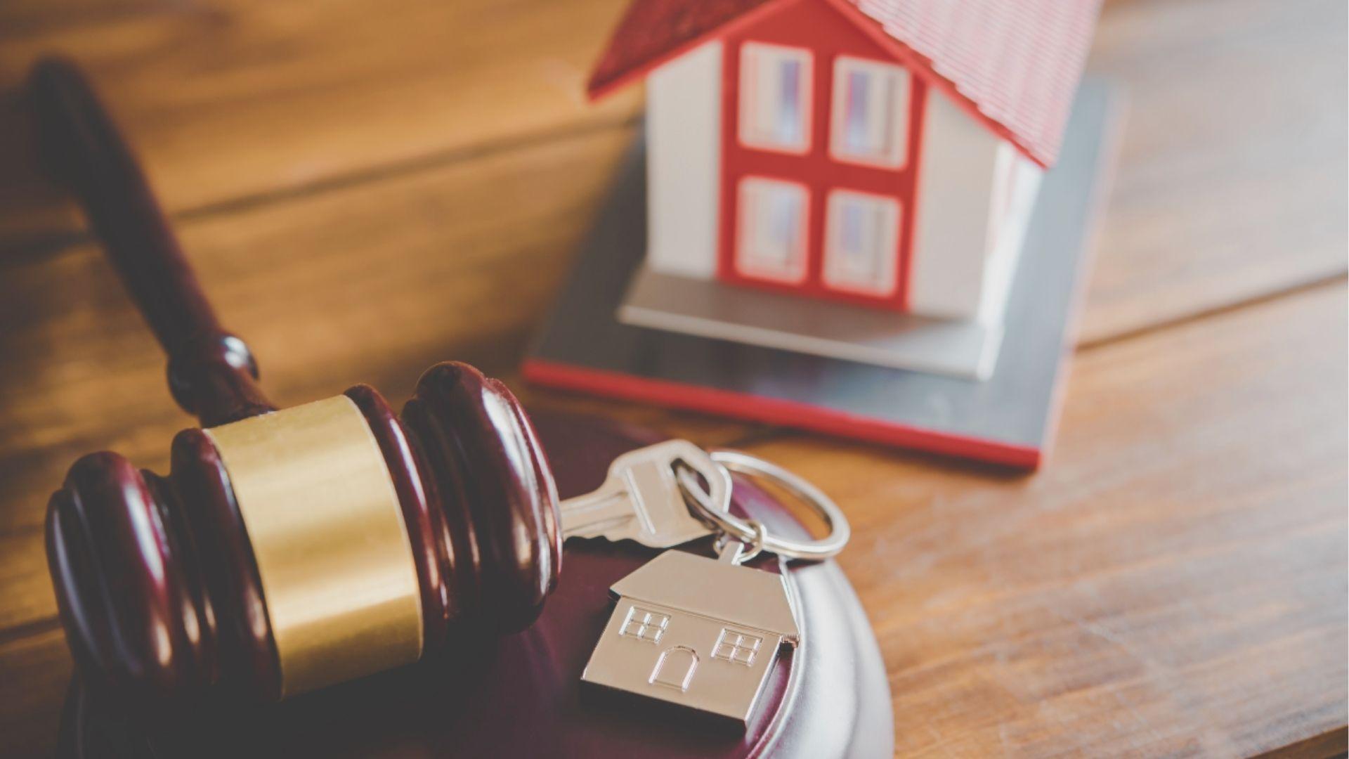 A judge’s gavel, house keys with a home-shaped keychain, and a red-roofed house model on a desk.