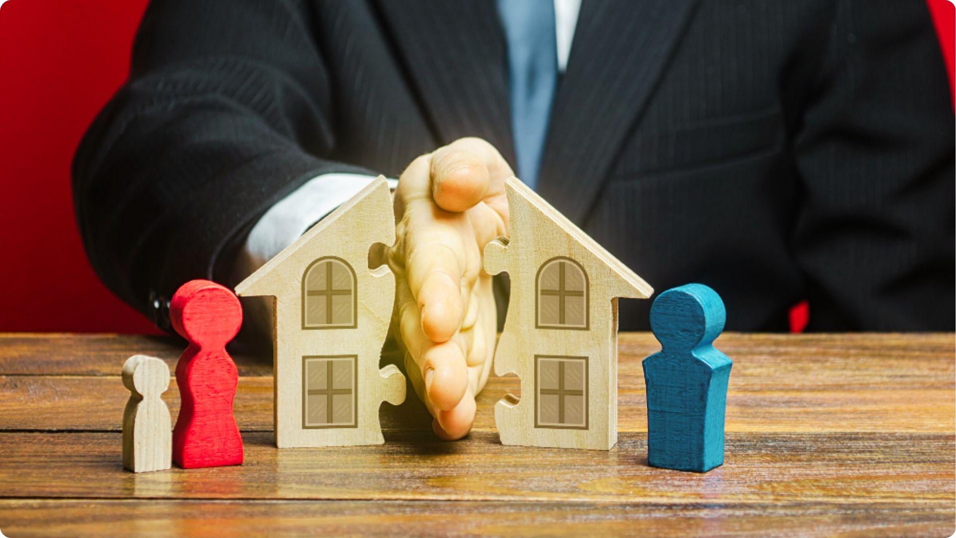 A person in a suit uses their hand to split a wooden house model between two wooden figures.