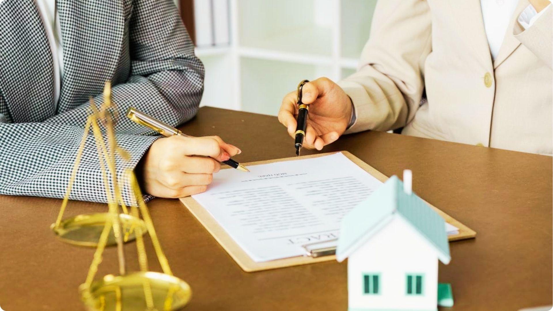 Two people signing a real estate contract with legal scales and a house model in the foreground.