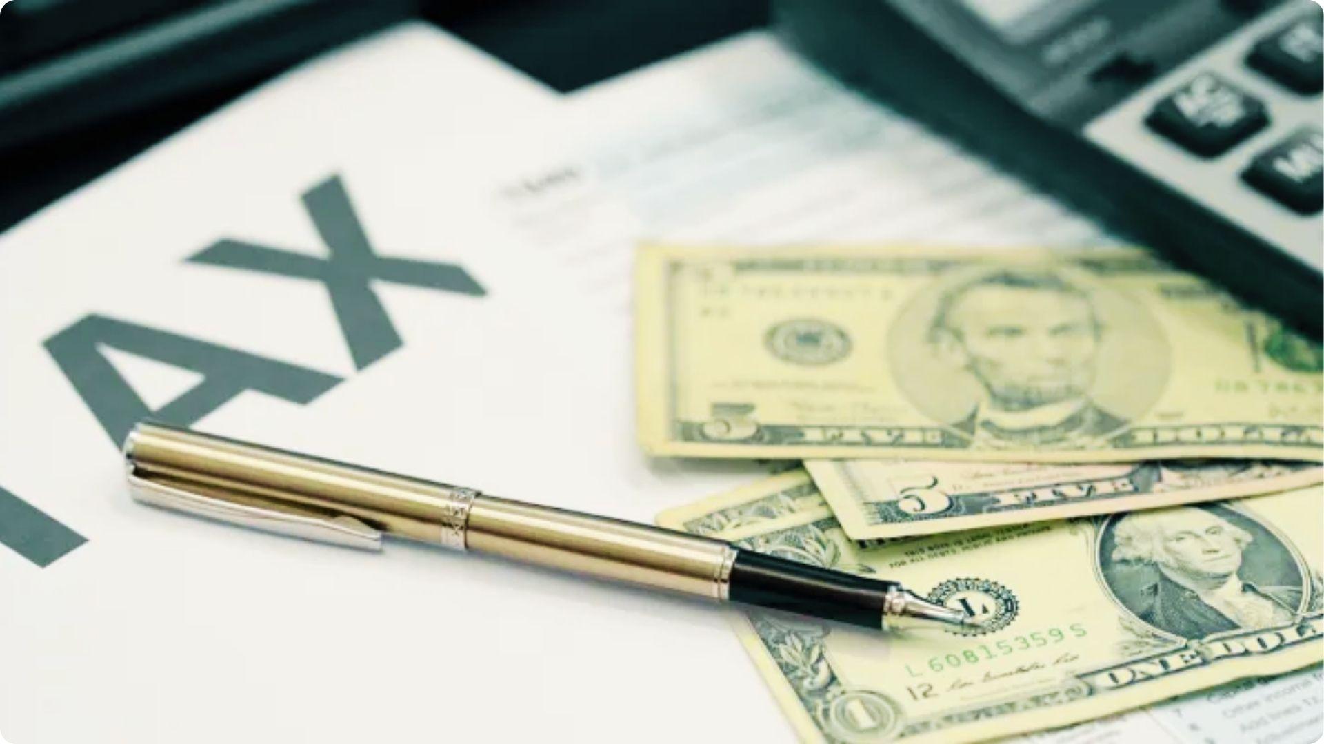 Pen, US dollar bills, and calculator rest on a document with large TAX text.