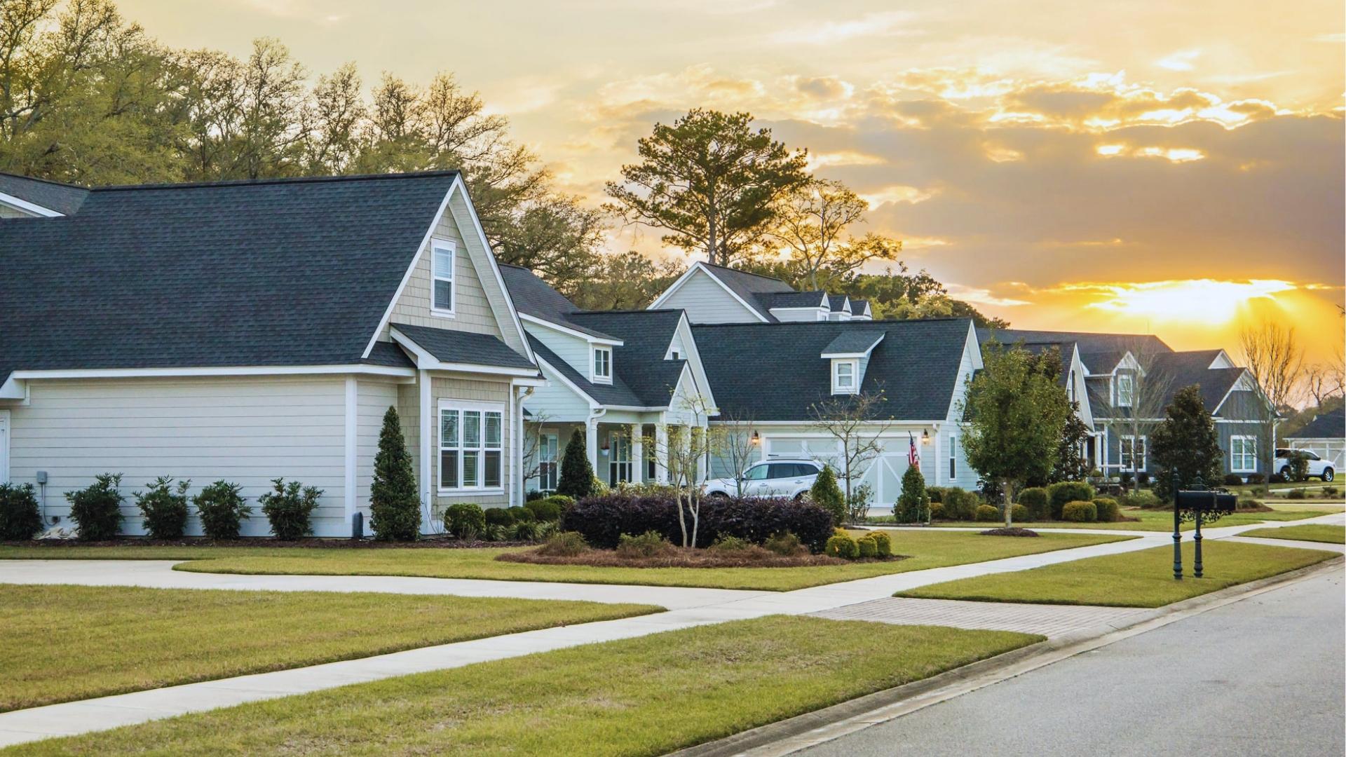 Single-story beige homes with dark roofs line street at golden sunset, with landscaped yards, sidewalks, and black mailboxes.