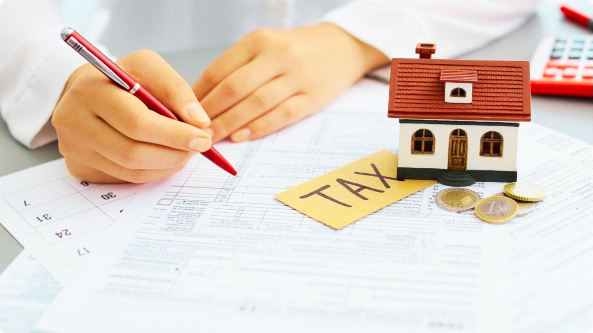 Hand with red pen fills out tax forms next to a model house, coins, and a TAX card.