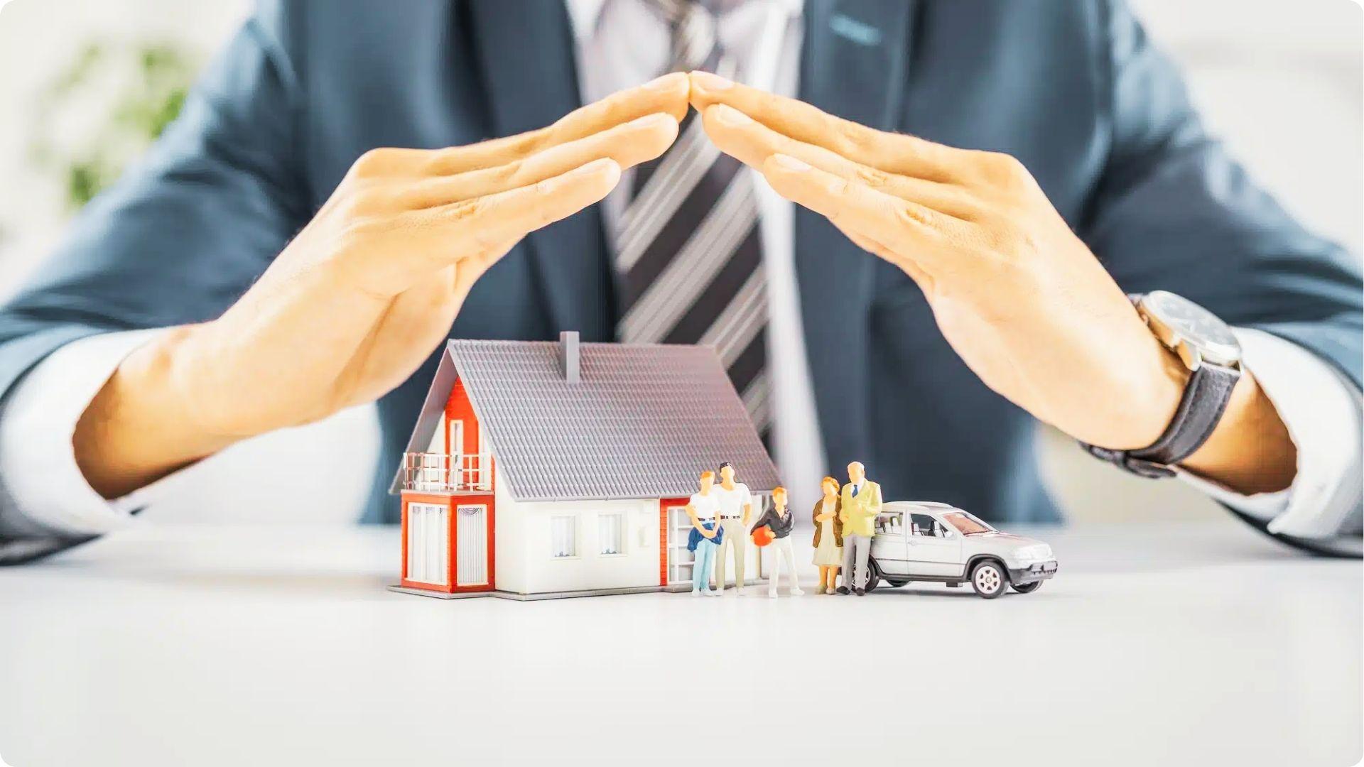 Businessman with hands forming a protective roof over a model home, family, and car.
