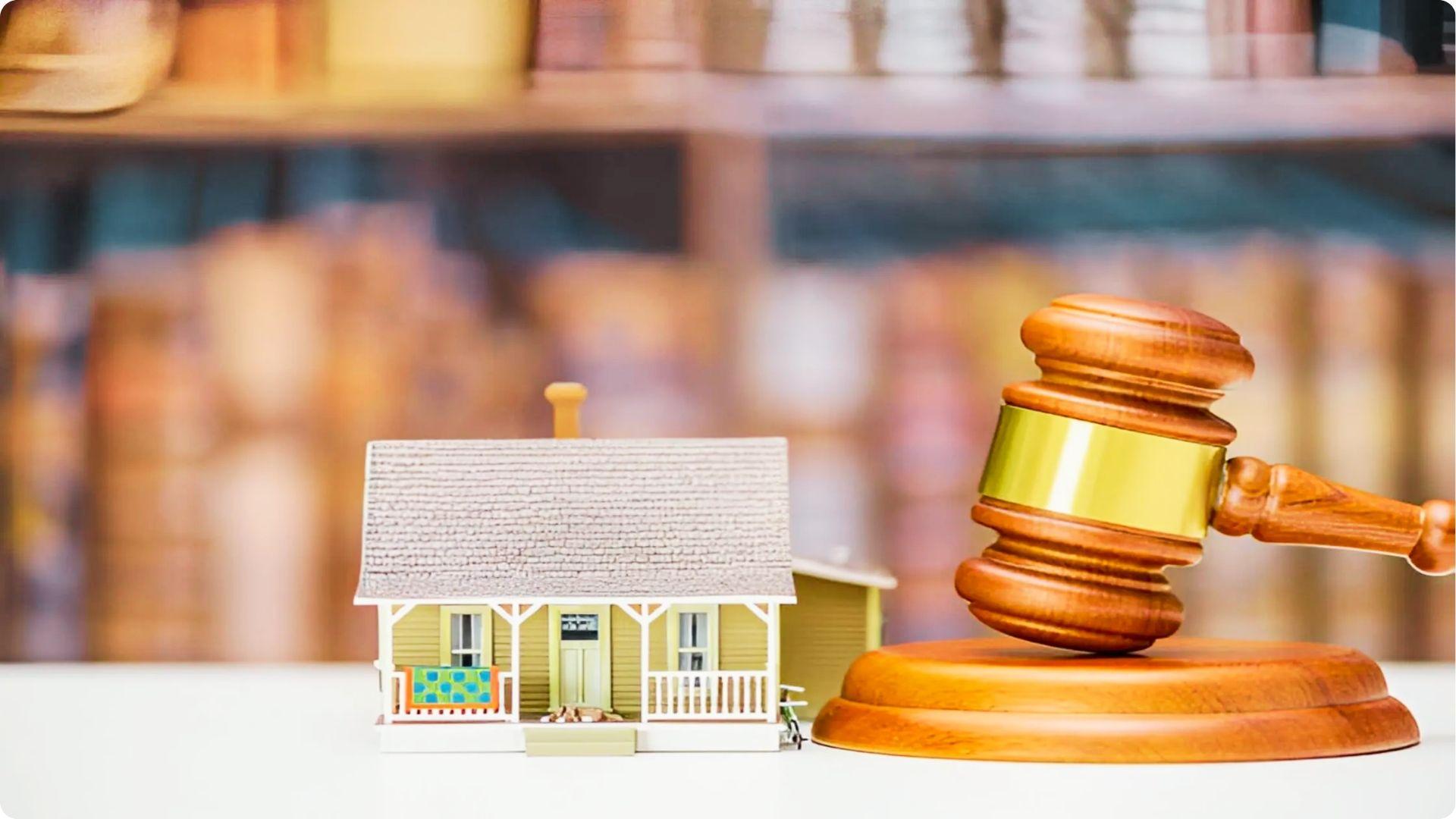 A wooden gavel rests next to a miniature house with law books in the background.