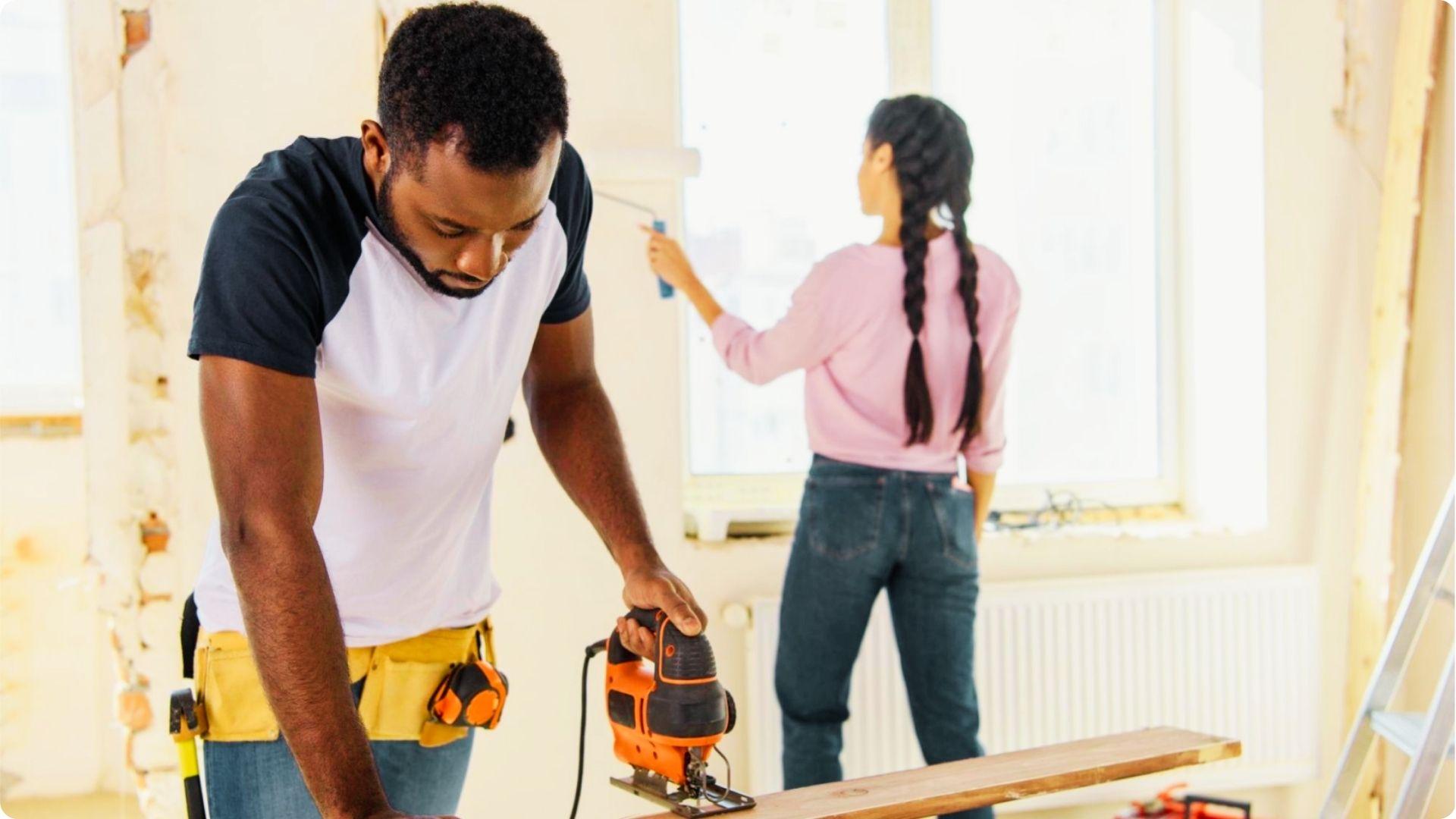 Man using a jigsaw and woman painting a window during a home renovation project.
