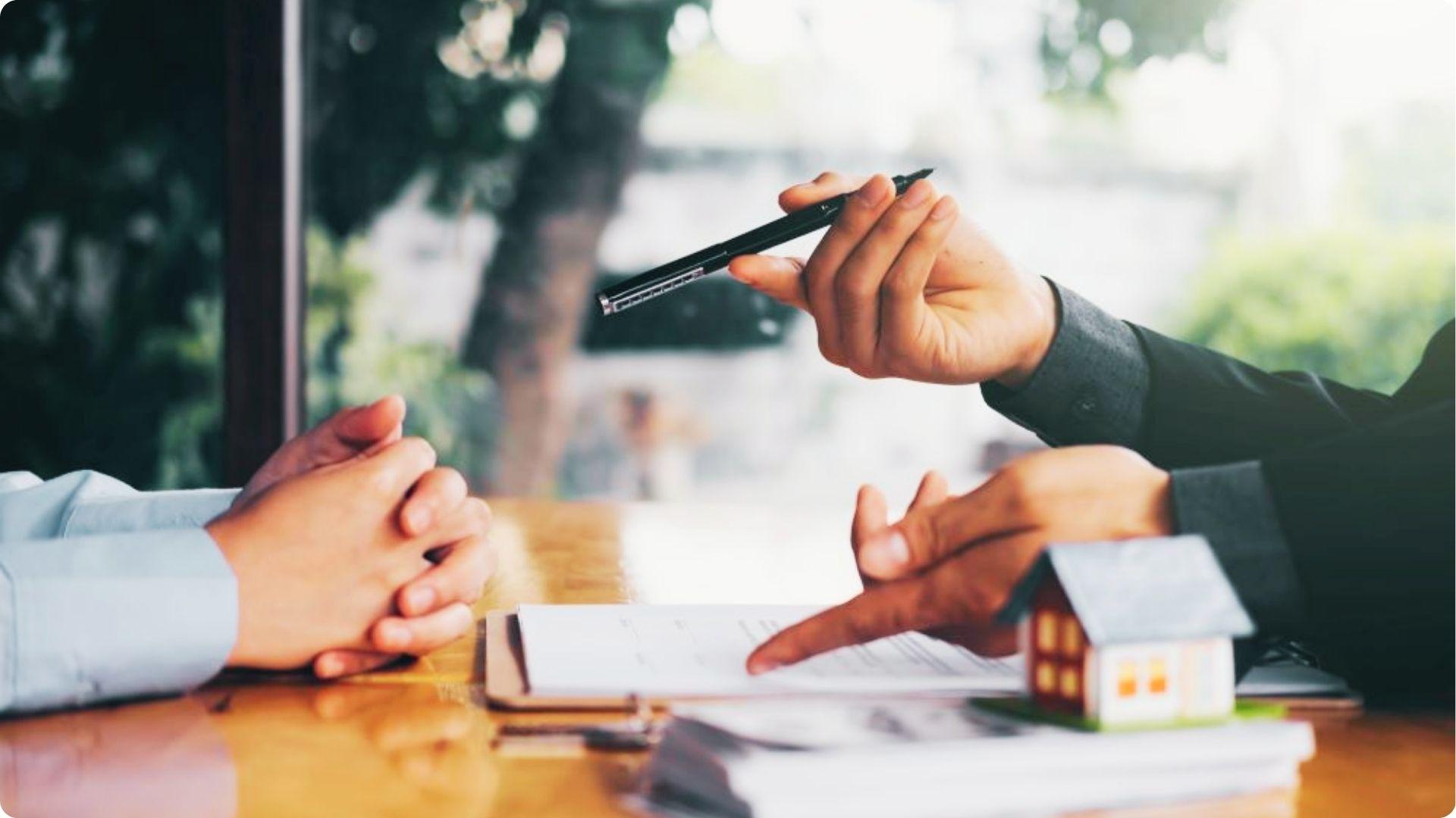 One person hands a pen to another over a contract with a small house model on the table.
