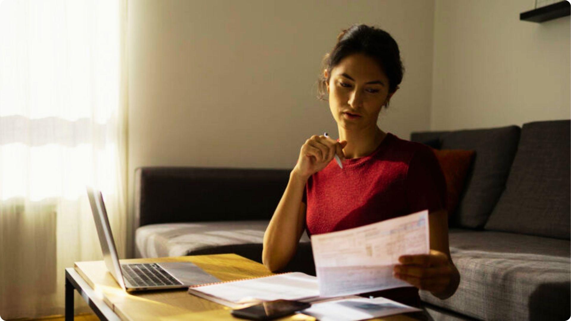 Woman in red shirt reviews tax paperwork at home with a laptop and notebook.