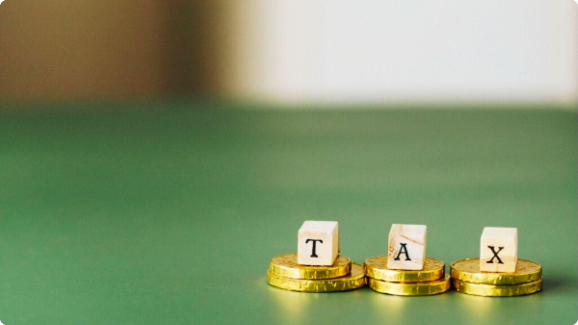 Wooden blocks spelling TAX sit on stacks of gold coins against a green background.