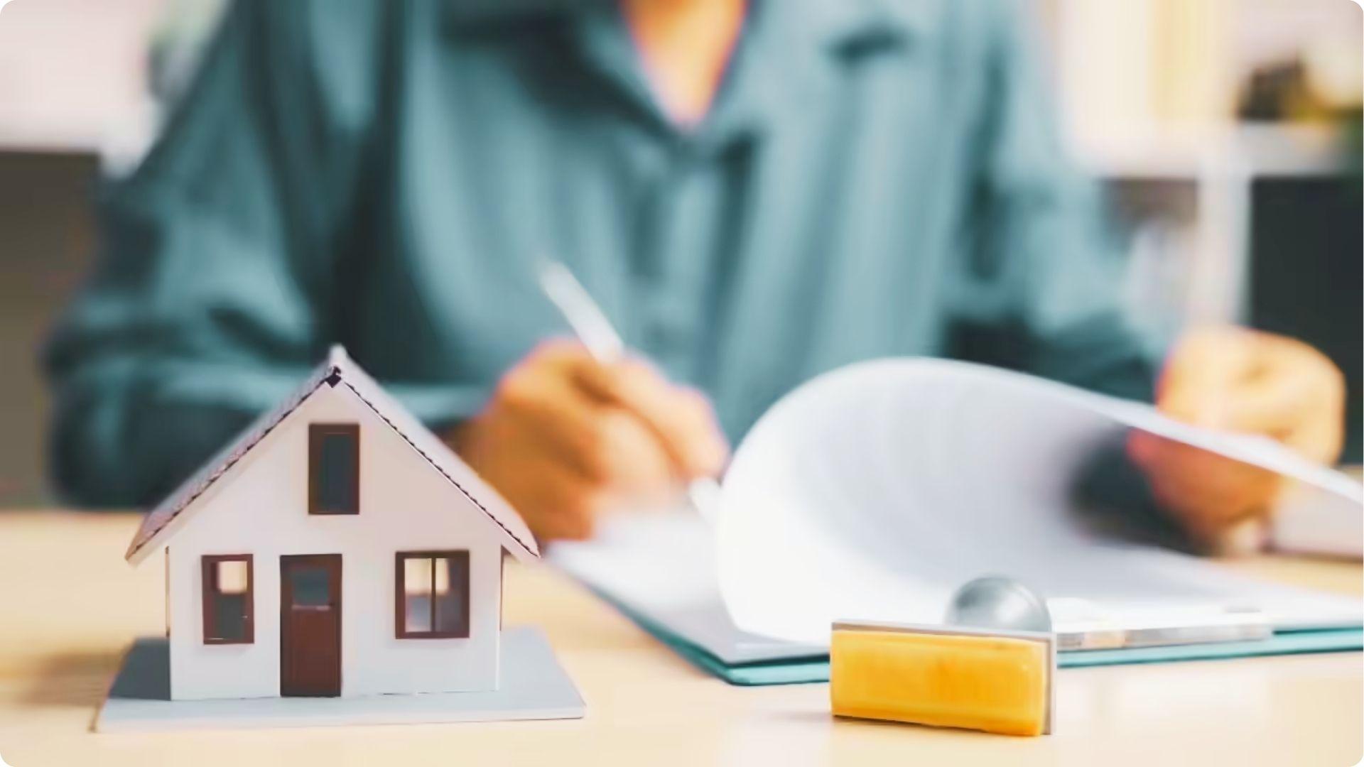 Close-up of a small house model beside signed documents and a stamp on a desk.
