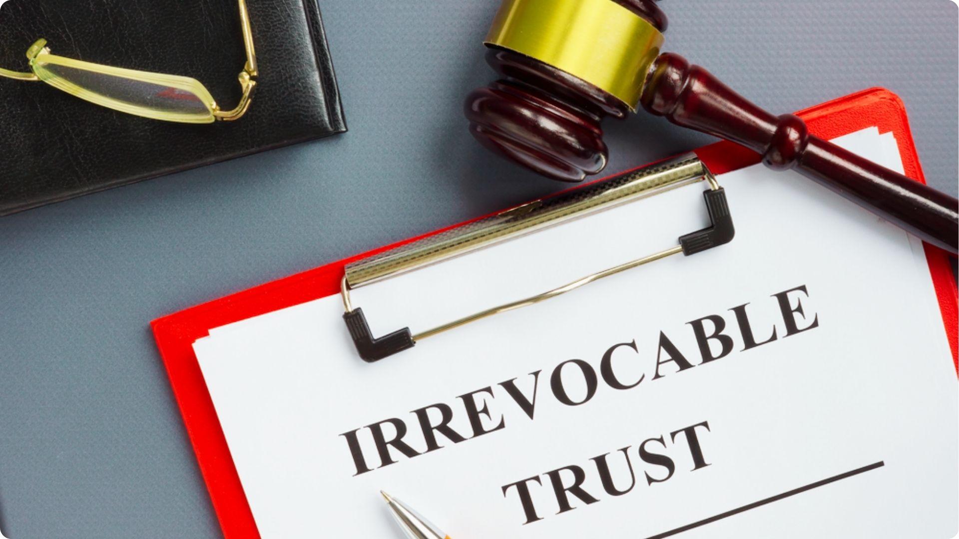 A clipboard labeled "Irrevocable Trust" lies next to a gavel and glasses on a gray desk.