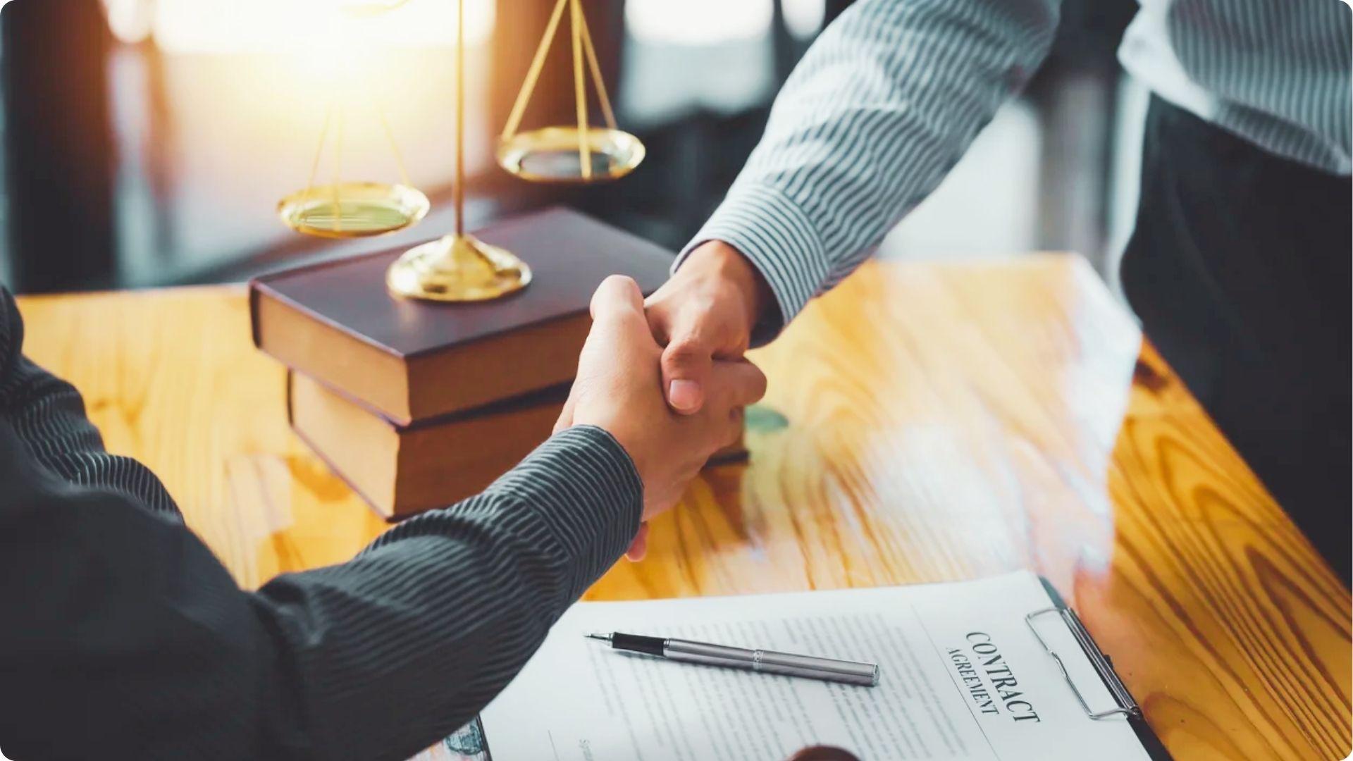 Two people shaking hands over a signed contract with scales of justice in the background.