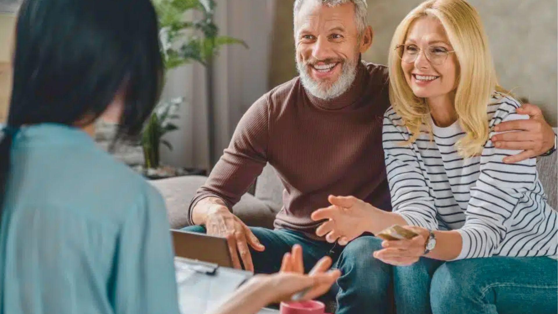 A smiling older couple sits with a financial advisor, reviewing documents in a living room.