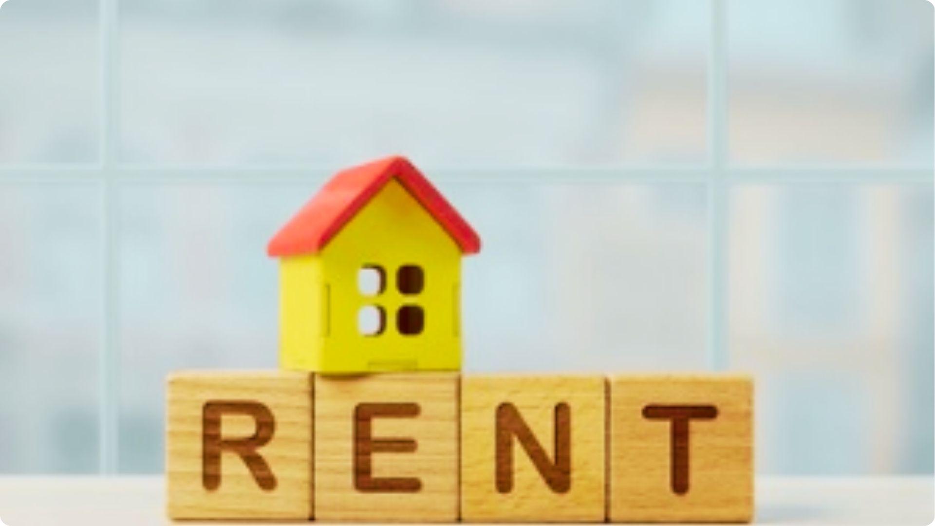 Wooden blocks spelling RENT support a small yellow house model with a red roof.