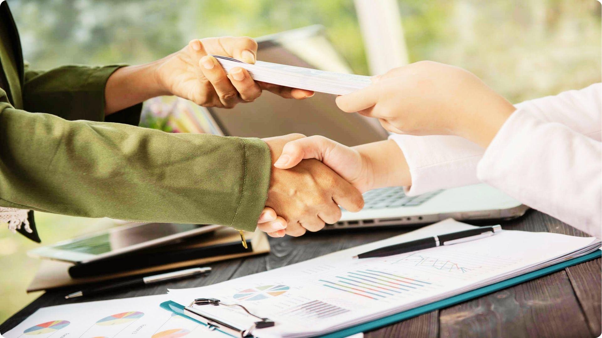 Hands exchanging money while shaking hands over financial charts and a laptop.