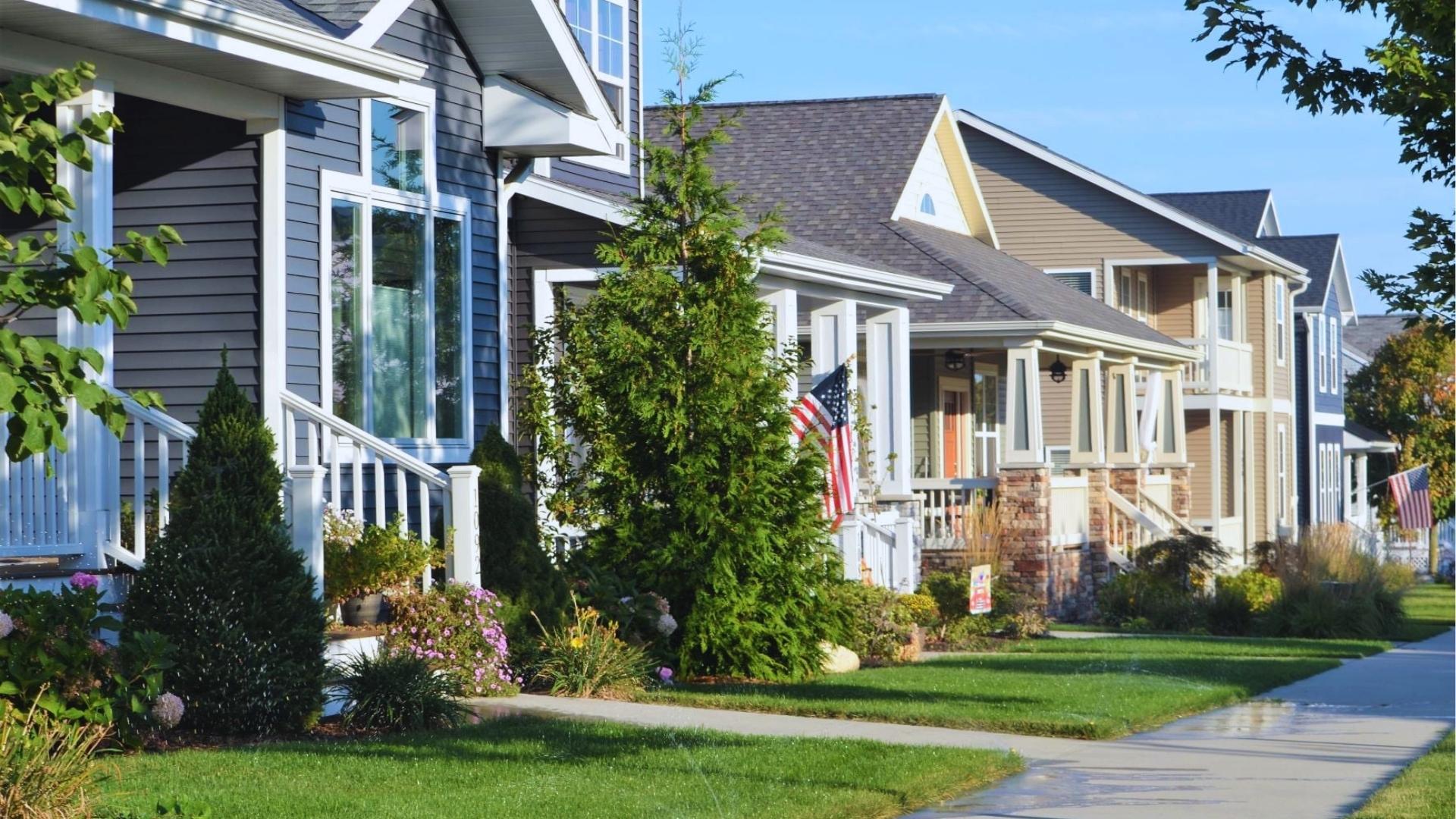 Row of suburban homes with dark blue and tan siding, white porches, American flags, and landscaped lawns along sidewalk.