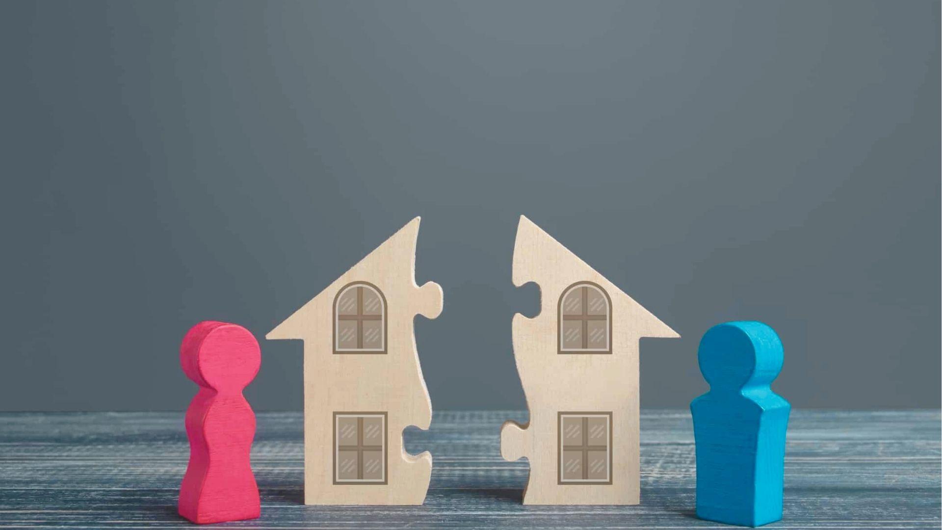 A wooden house cut into two puzzle pieces sits between pink and blue figurines on a table.