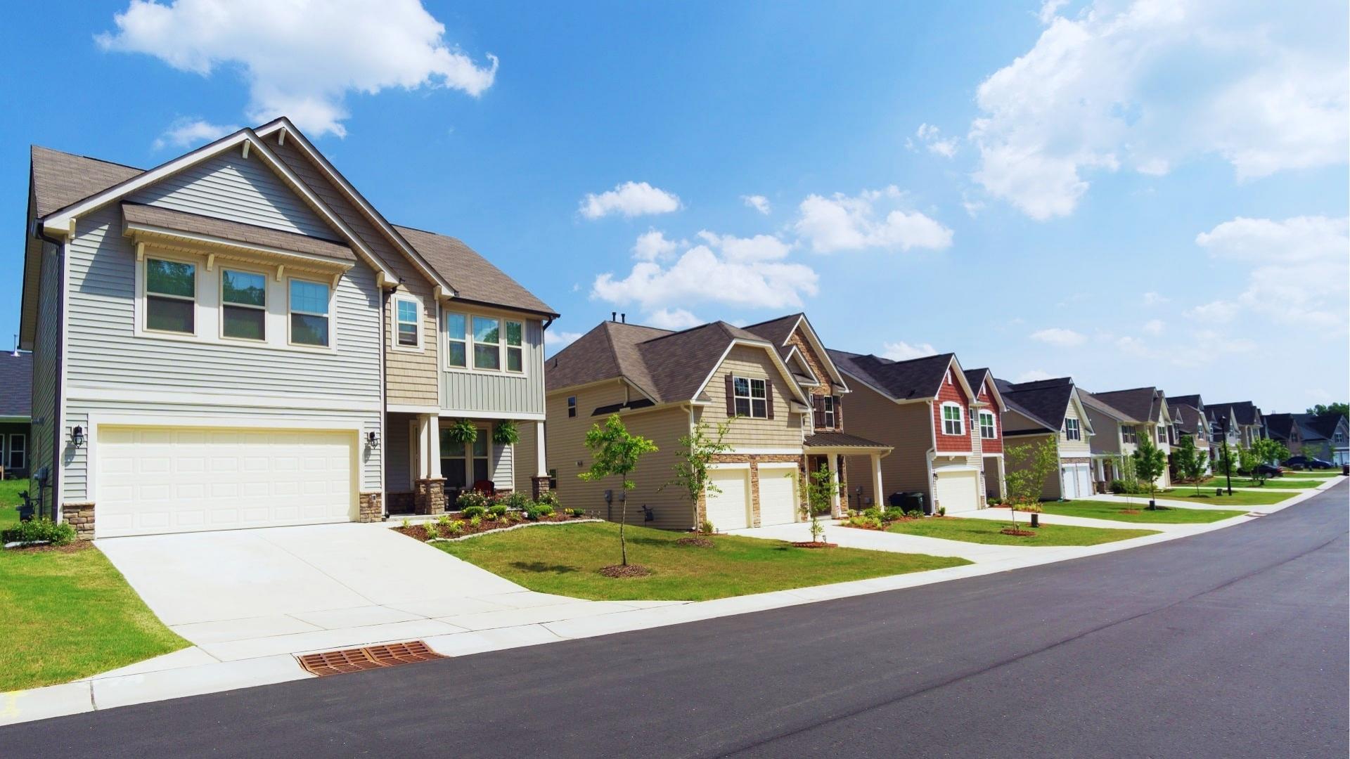 Modern two-story houses with attached garages line a curved suburban street under blue sky with green lawns and storm drain.