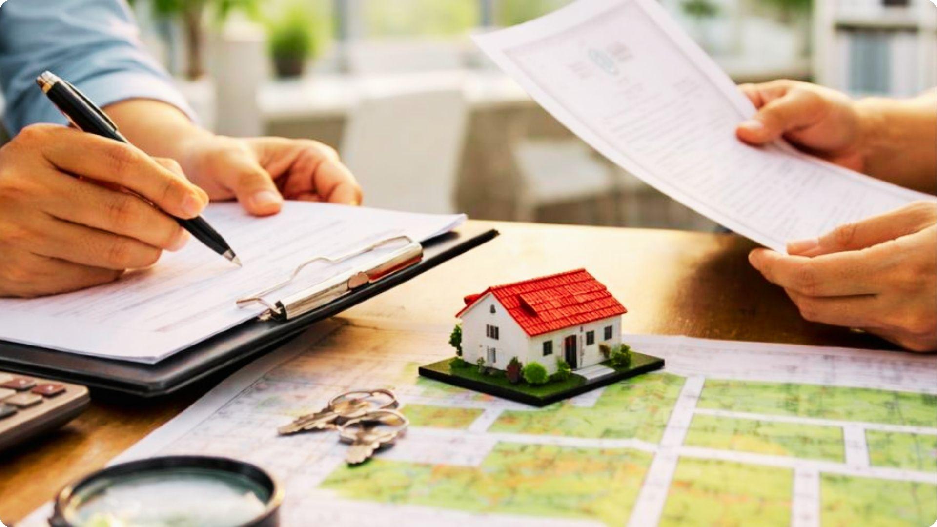 Two people review real estate documents at a desk with a model house, keys, and property map.