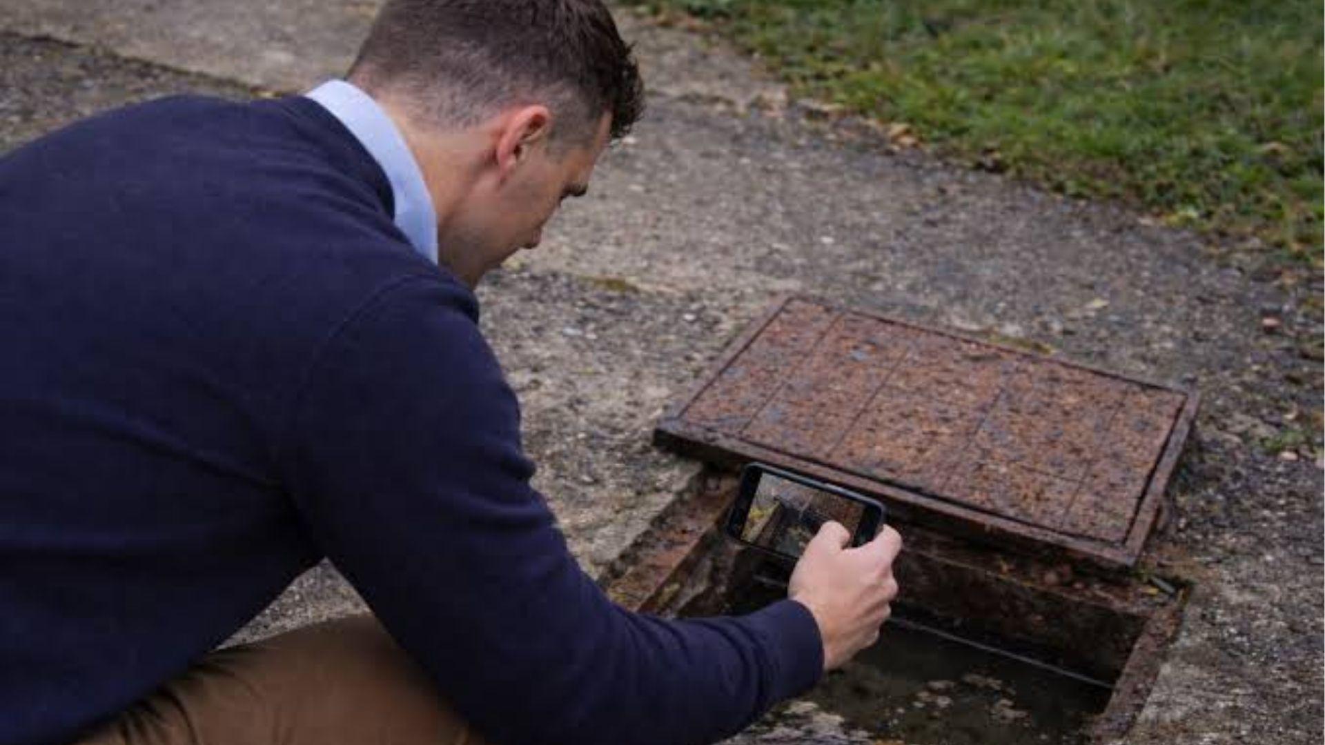 Surveyor inspecting an open utility access cover with a smartphone outdoors.