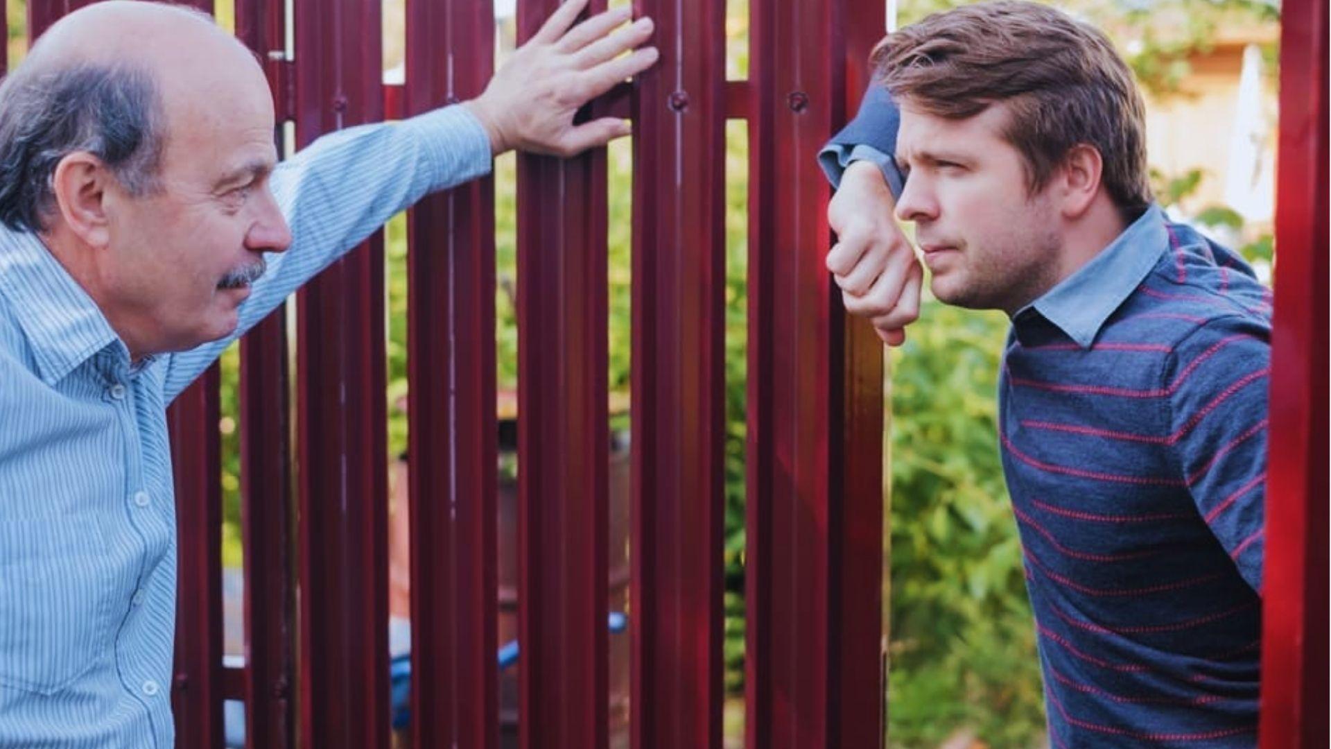 Two men in a tense argument facing each other through a red metal fence outdoors.