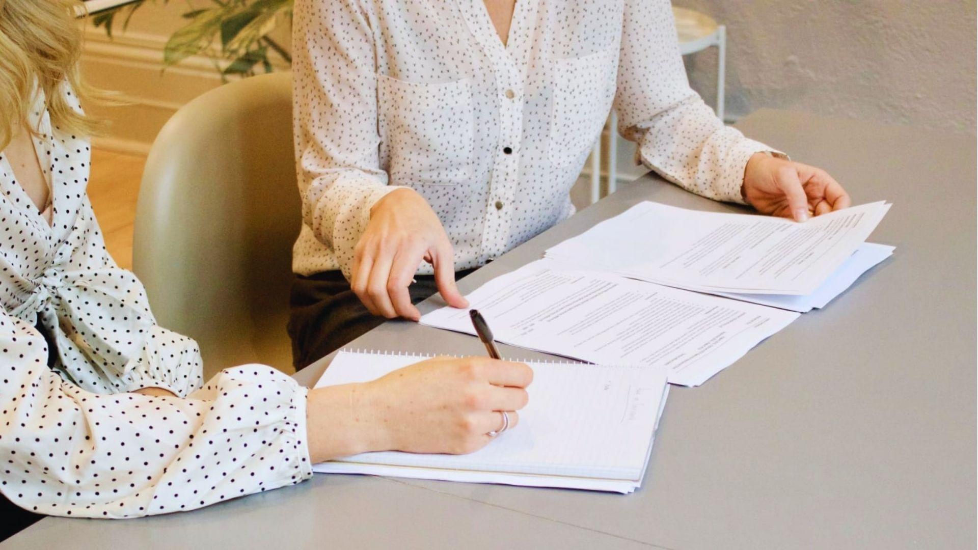 Two women at a gray table reviewing printed documents, one pointing at paperwork while the other writes notes in a notebook with a pen.
