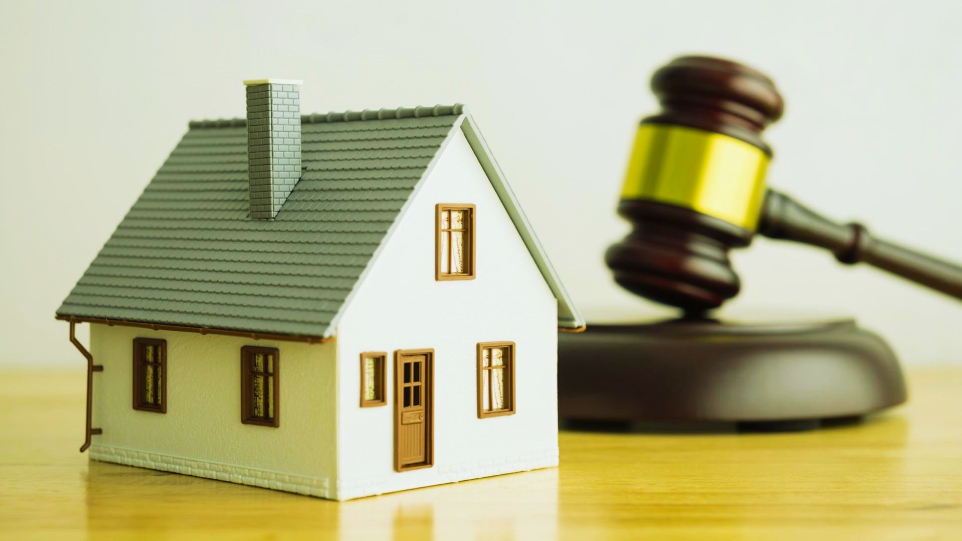 A miniature model house resting on a wooden surface next to a wooden judge's gavel.
