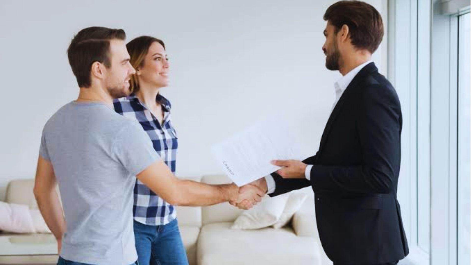 Couple shaking hands with suited agent holding contract document in bright living room.